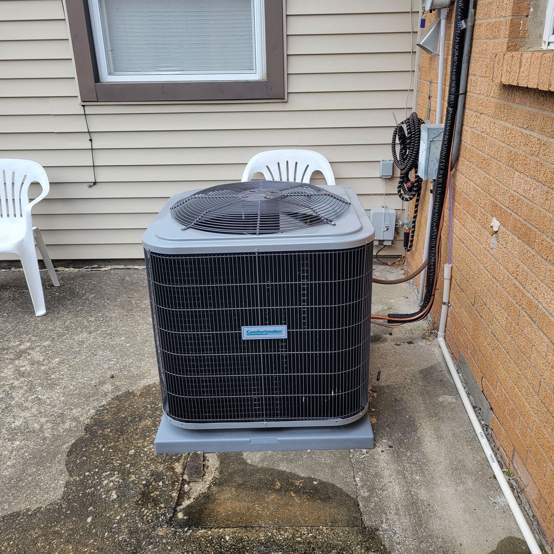 Outdoor air conditioner unit beside a house wall on a concrete pad, with patio chairs nearby.