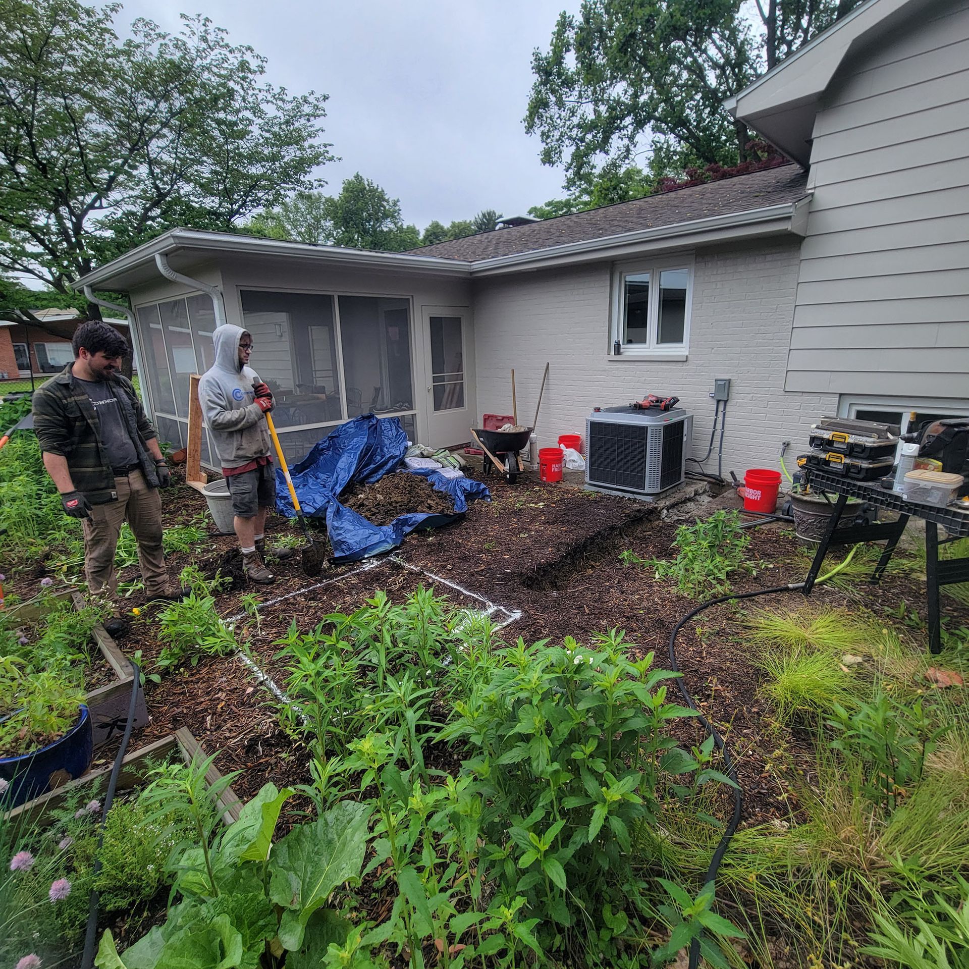 Two workers clearing storm debris beside a house, with piles of branches, a tarp, and yard tools.