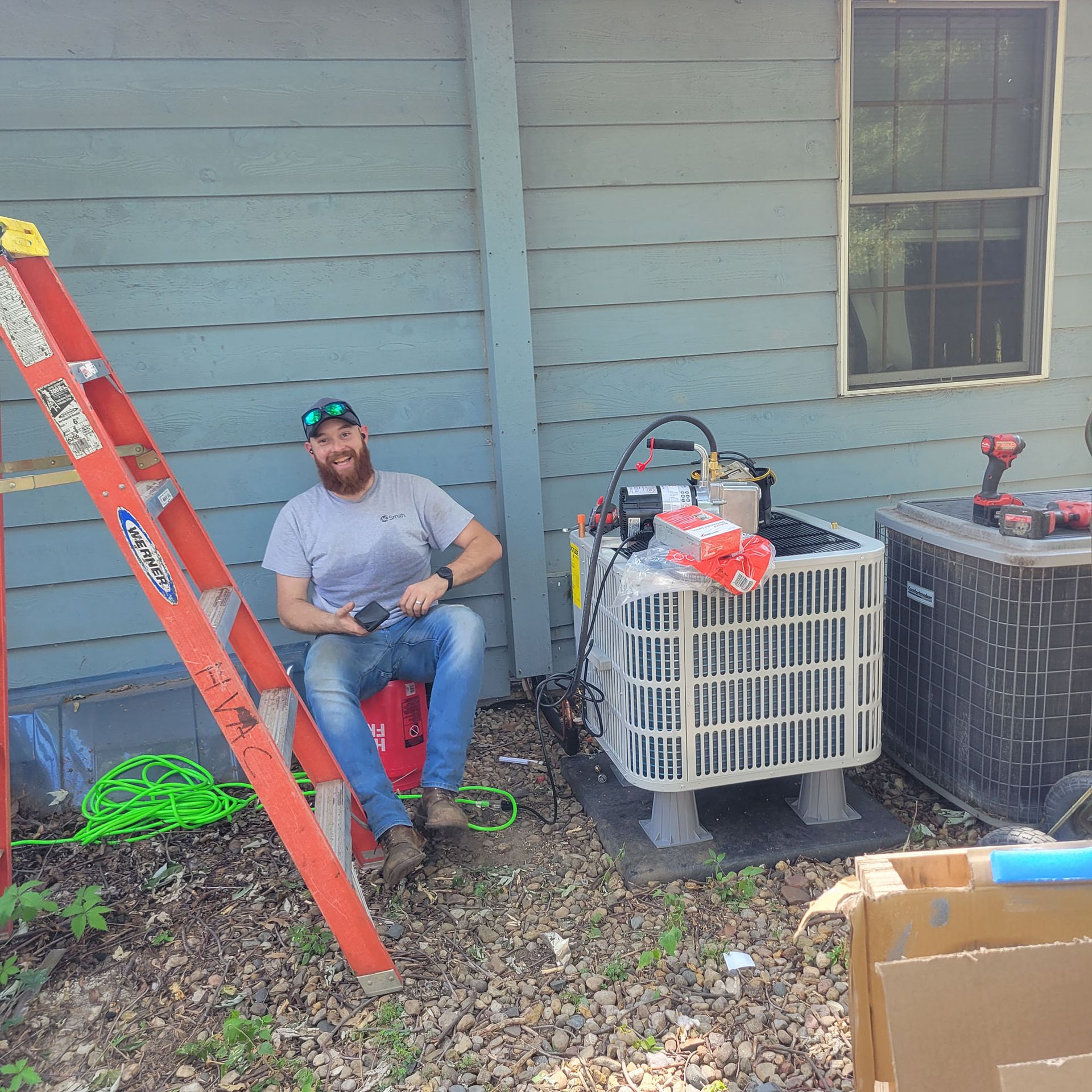 Man sitting on a red stool by a house wall near a ladder and two outdoor HVAC units