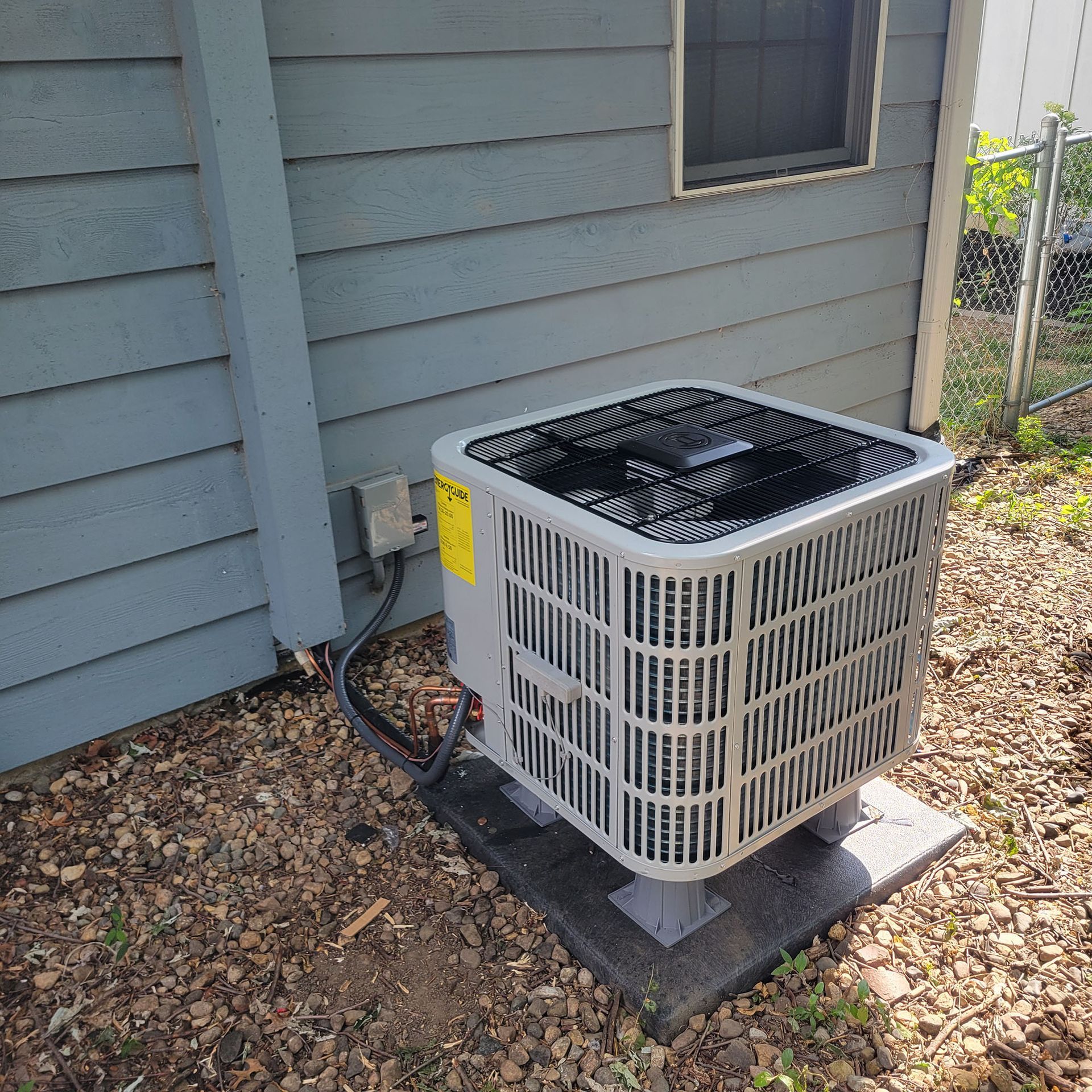 Outdoor air conditioner unit beside a gray house wall, sitting on a concrete pad.