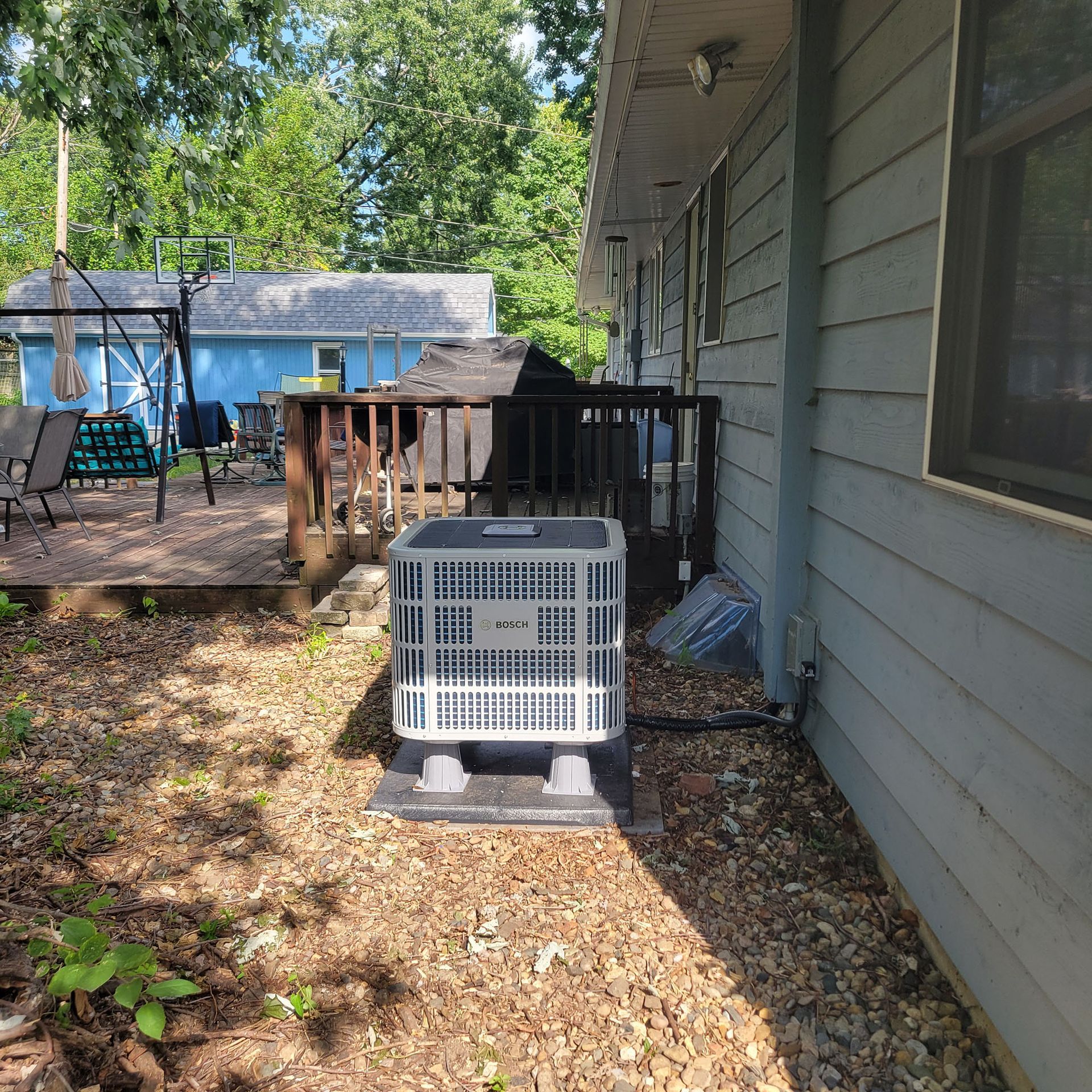 Backyard patio beside gray house, with white plastic crate on a black base and trees in the background