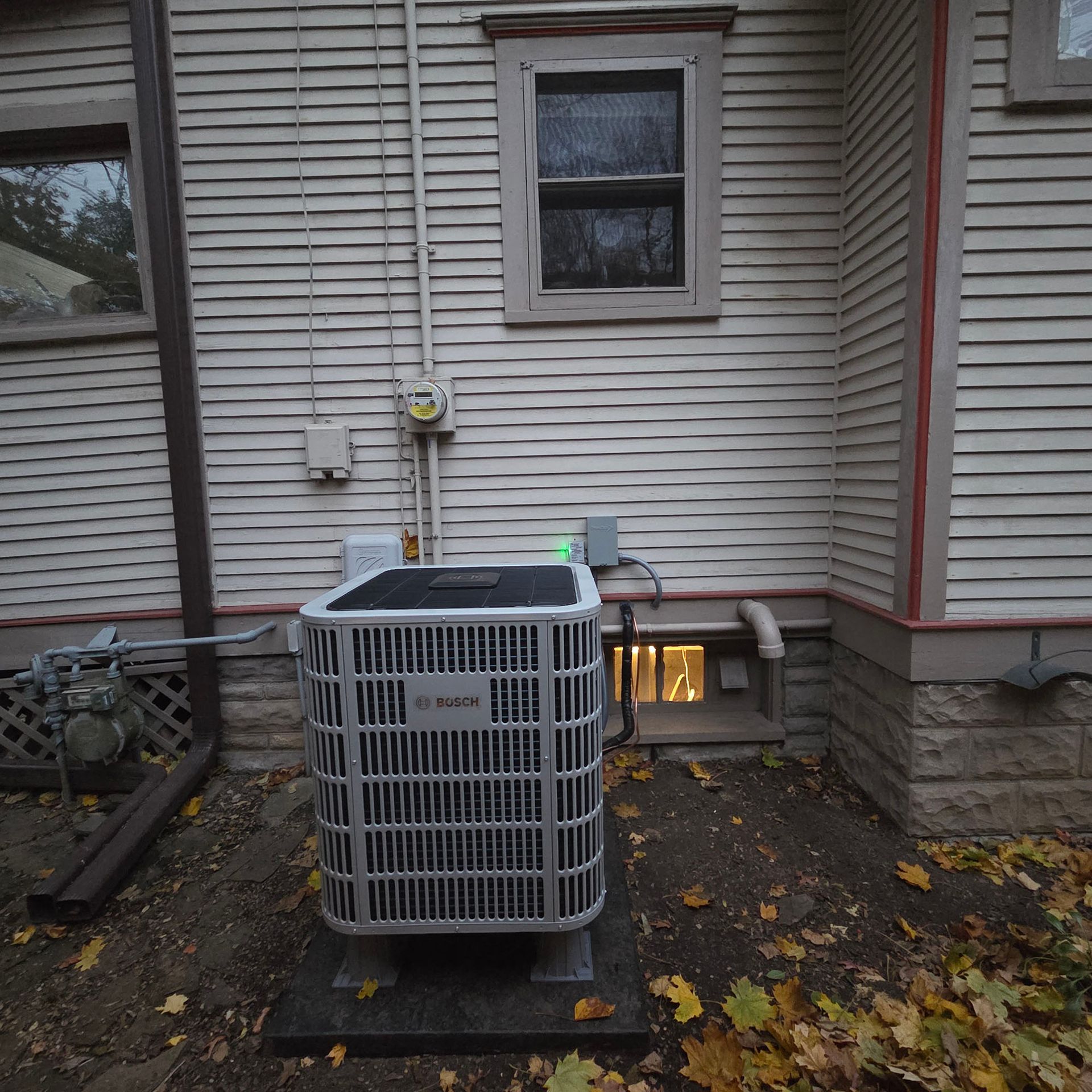 Outdoor HVAC unit beside a house wall with basement window and autumn leaves on the ground