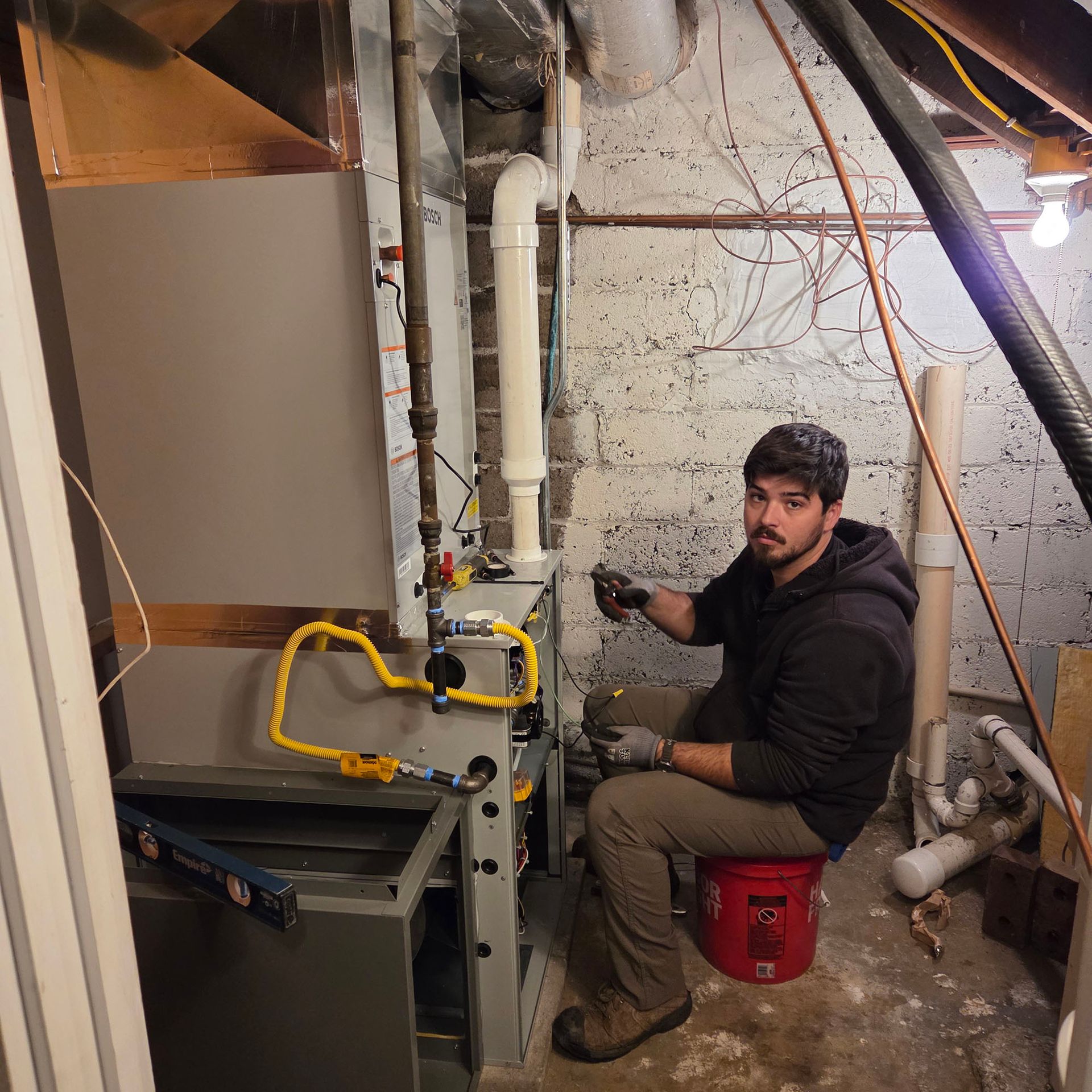 Man in a basement utility room working on furnace pipes beside a red bucket