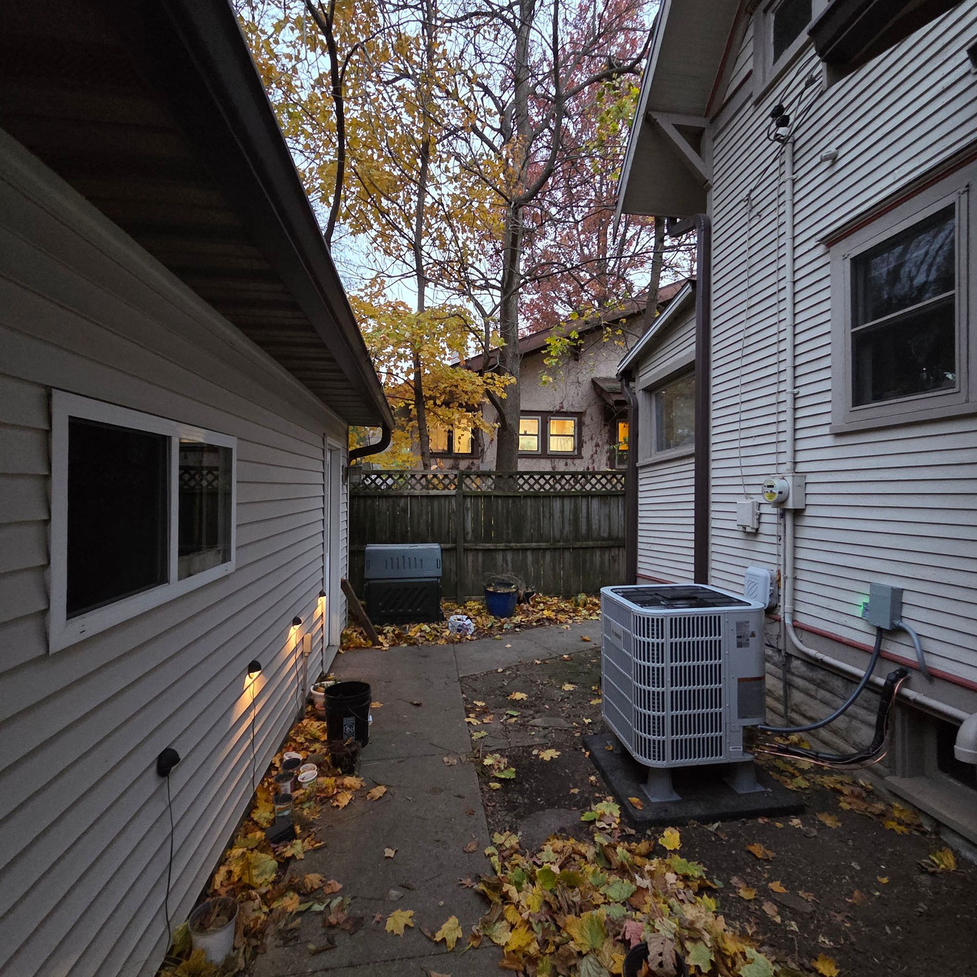 Narrow side yard between two houses with fallen leaves, HVAC unit, and a wooden fence at dusk.