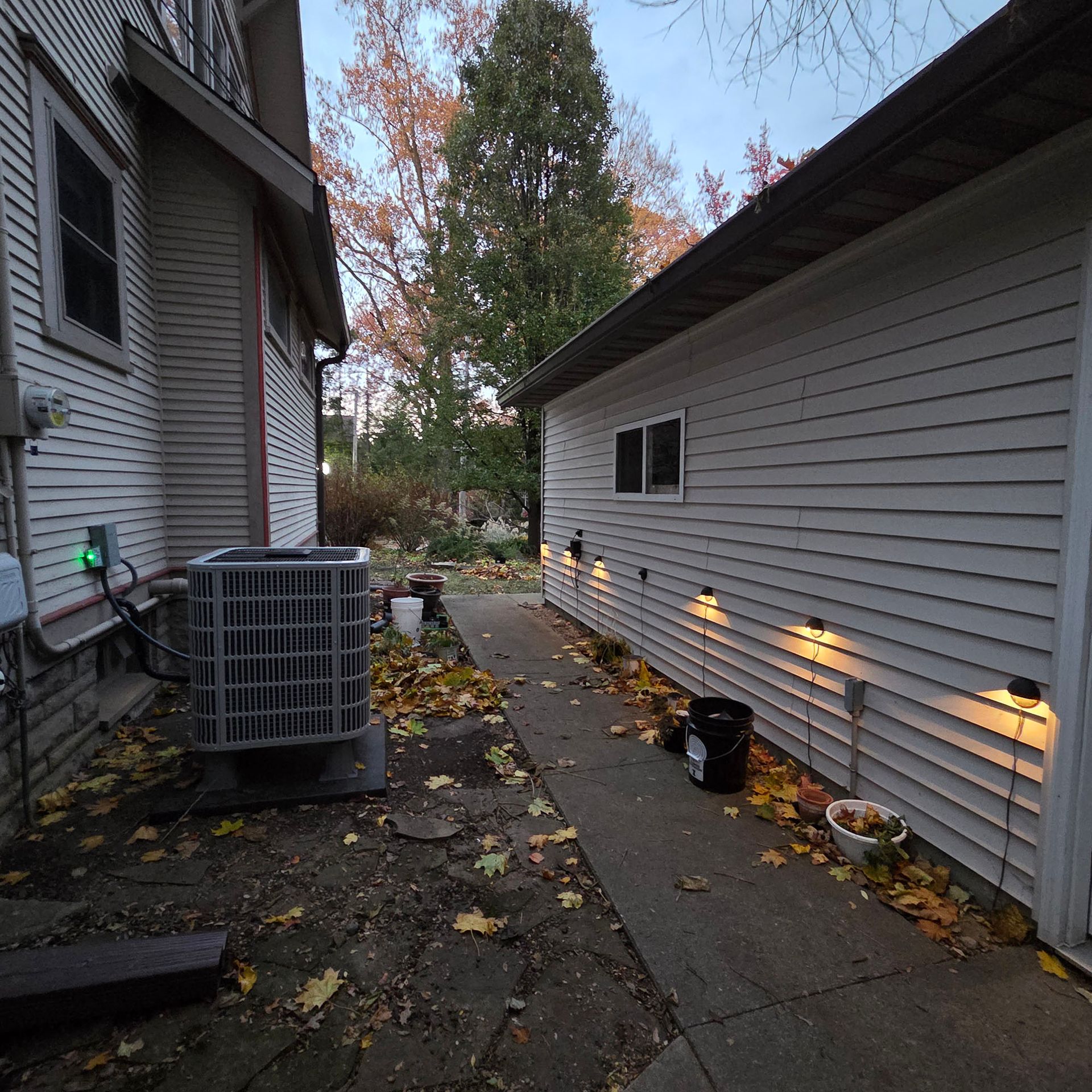 Narrow side yard between two houses with an AC unit, concrete path, scattered leaves, and lit wall lights