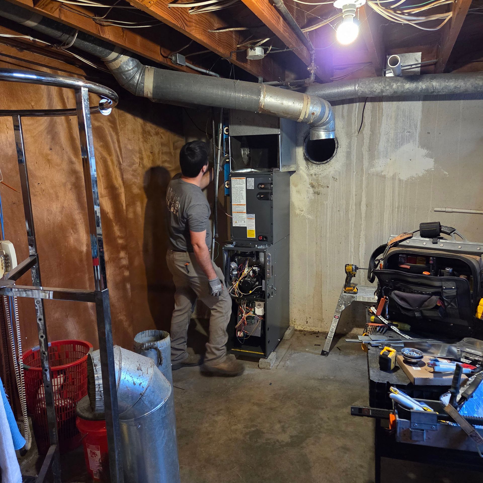 Person inspecting a furnace in an unfinished basement with exposed ductwork and tools nearby