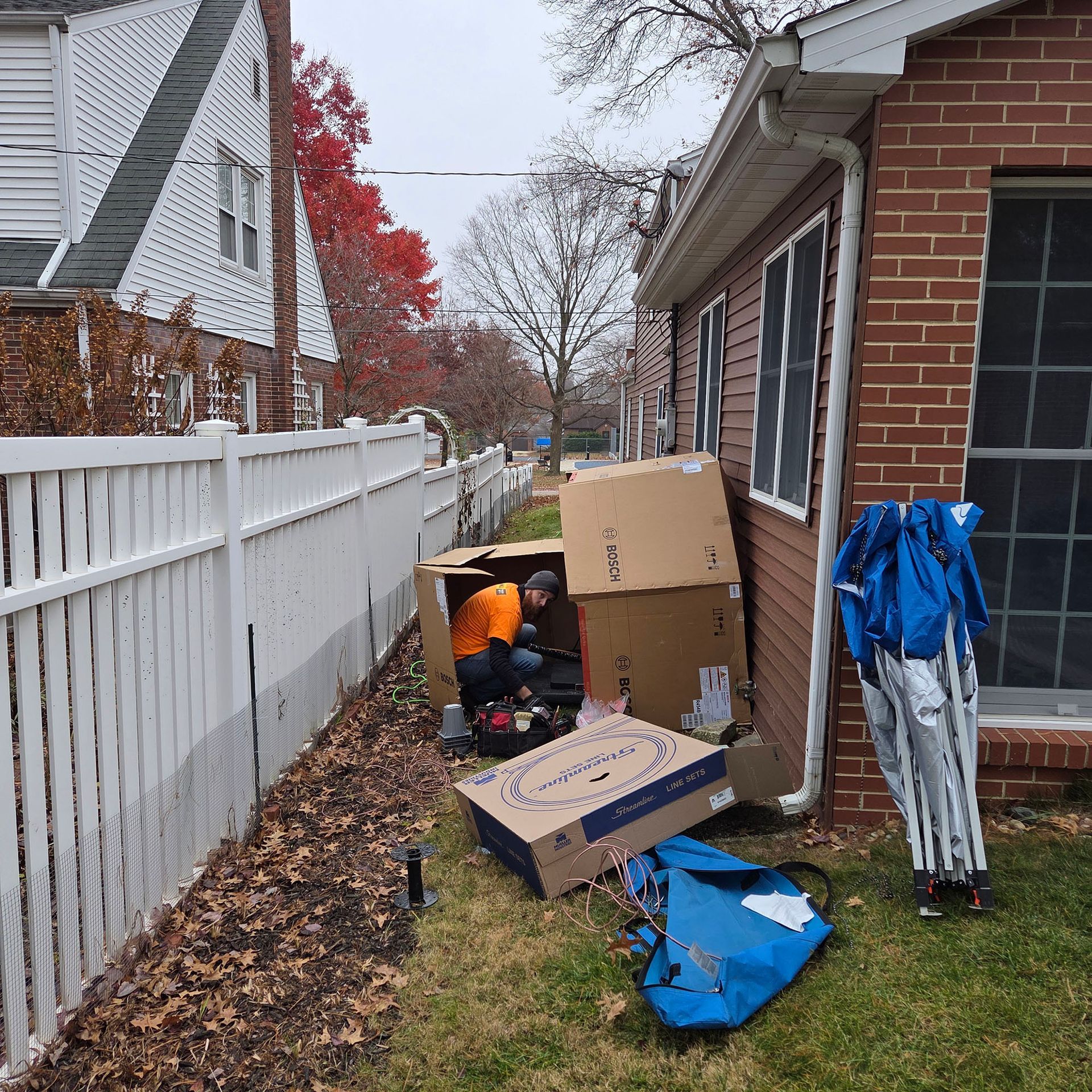 Cluttered side yard beside a brick house and white fence, with boxes, blue tarp, and a folded striped chair.