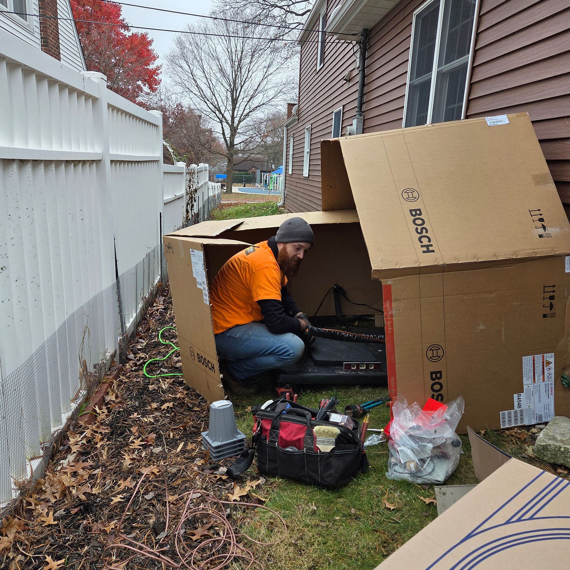 Person kneeling inside a cardboard box outdoors beside a house, surrounded by tools and bags.