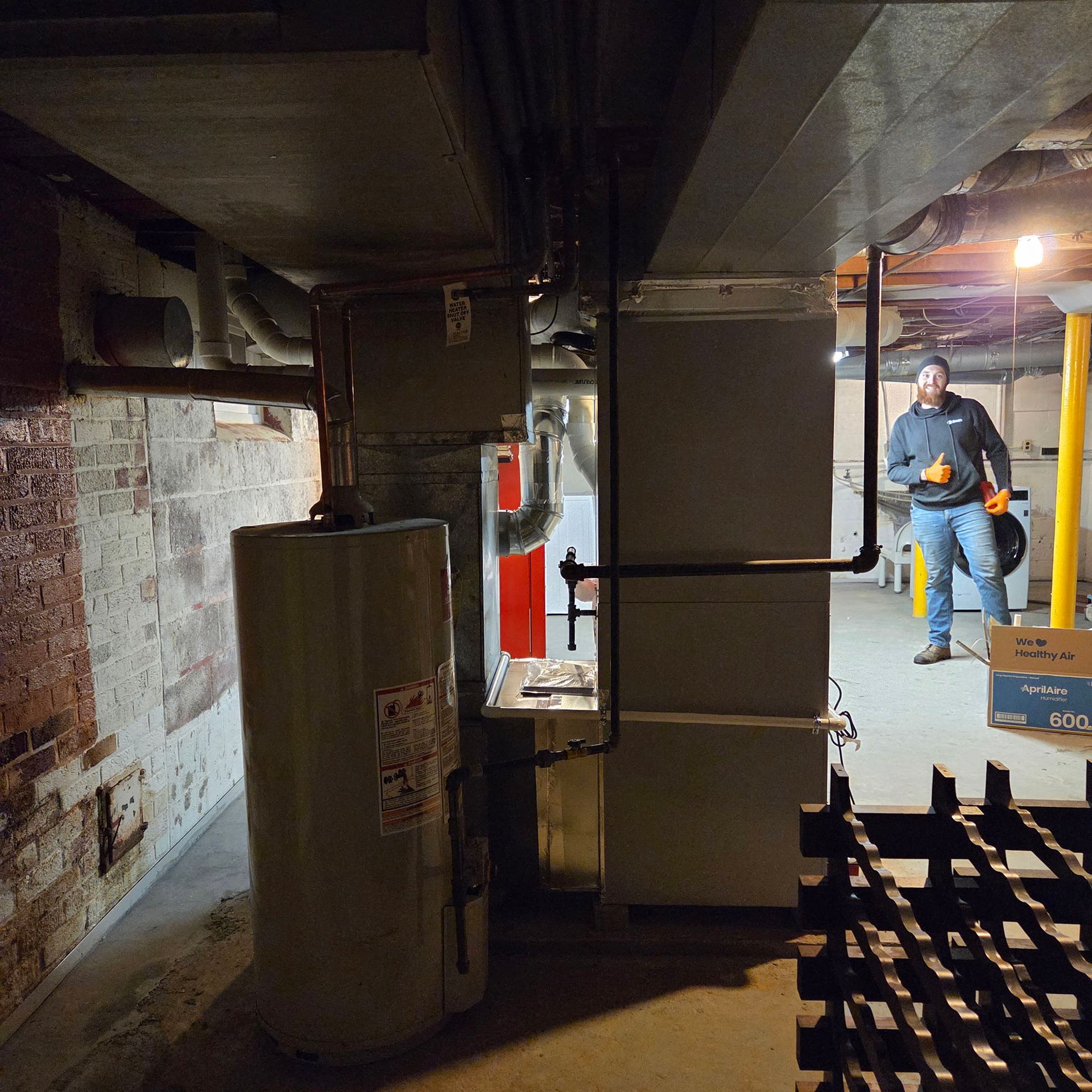 Basement utility room with pipes and equipment; worker in orange vest stands near stacked materials.