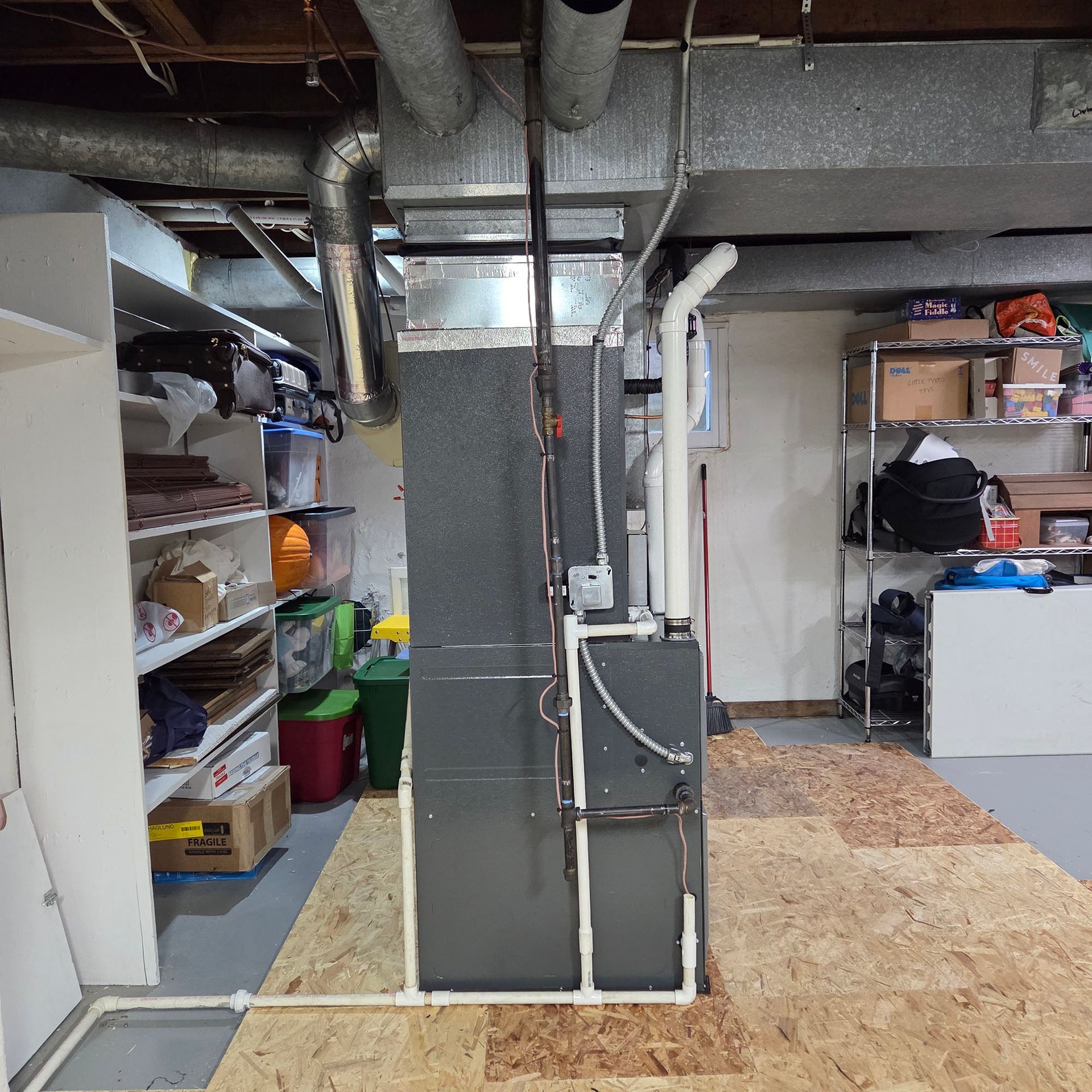 Basement utility room with exposed ducts, shelving, and storage boxes on plywood flooring.