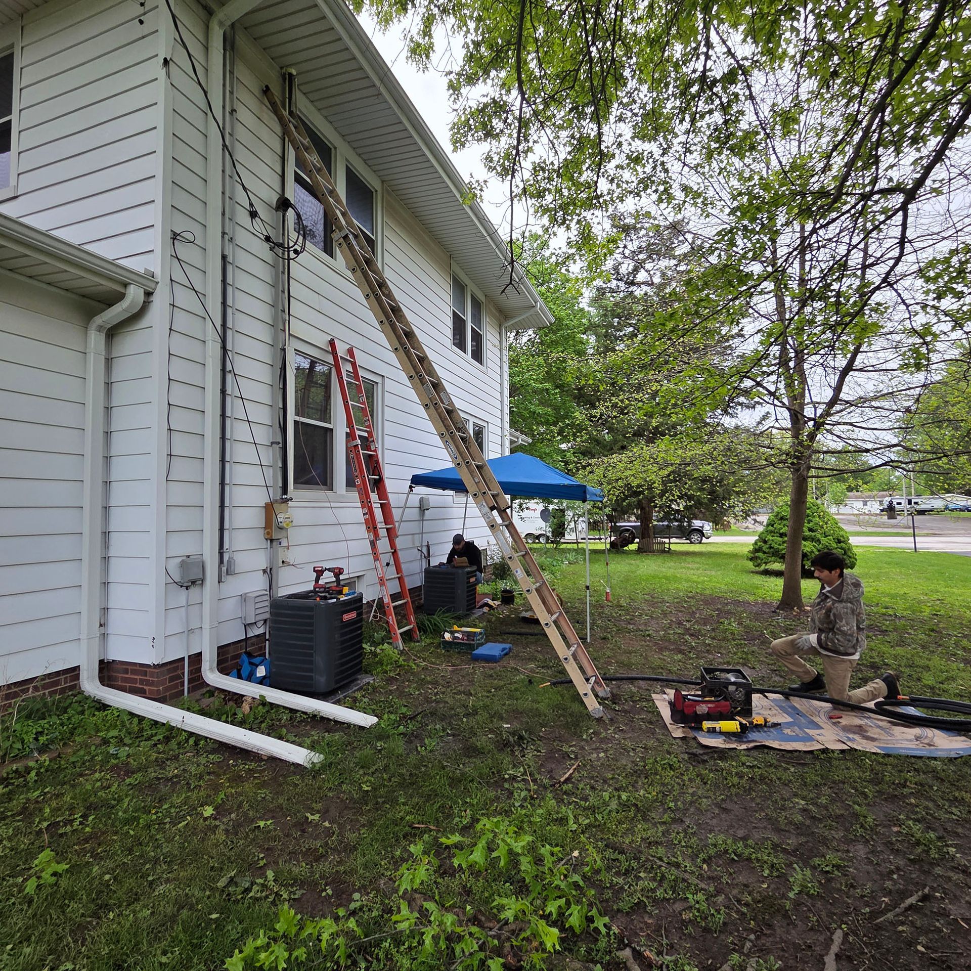 Workers on ladders repair a white house siding near a yard with tools and a blue umbrella.