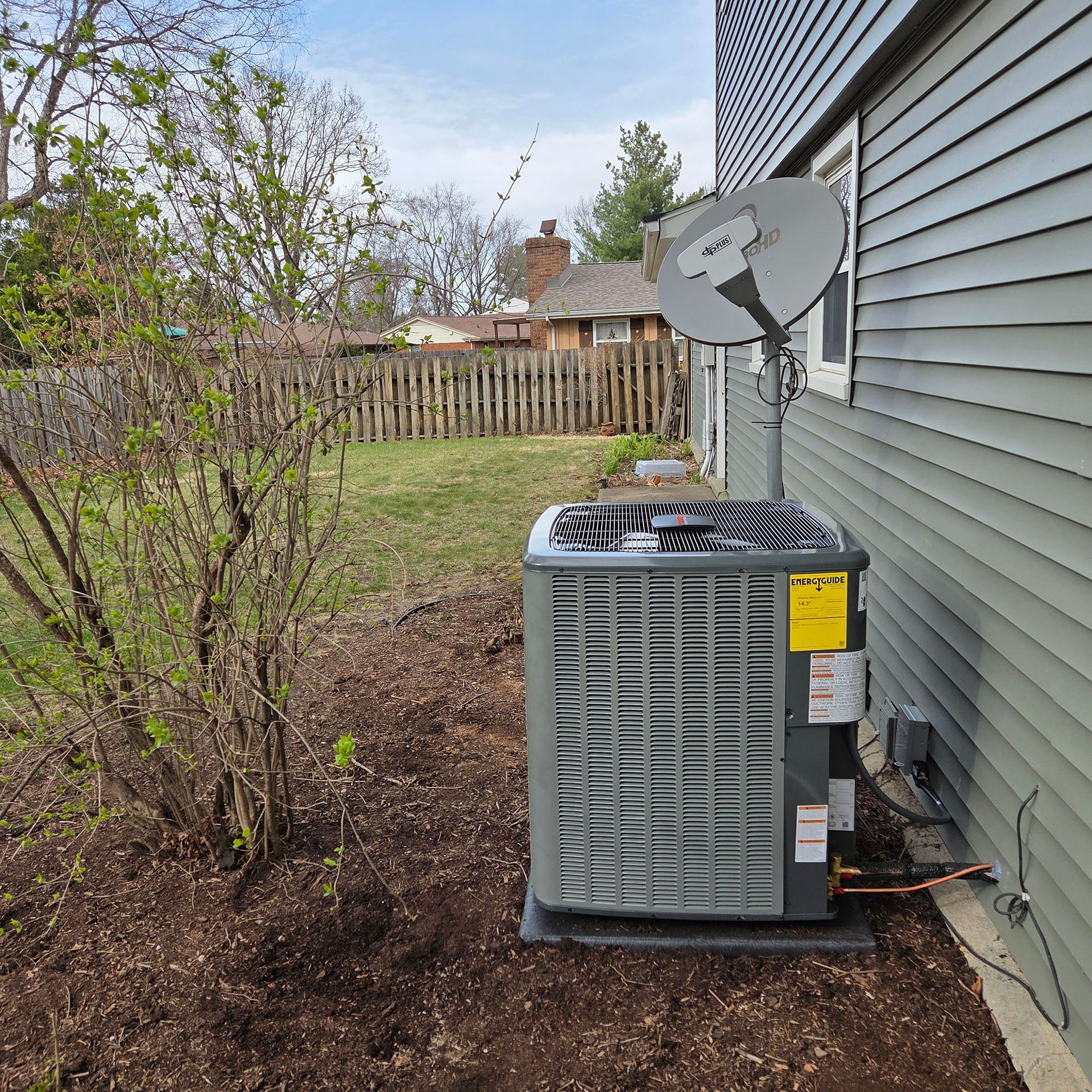 Outdoor HVAC unit beside a house wall with a satellite dish, yard, and wooden fence in the background