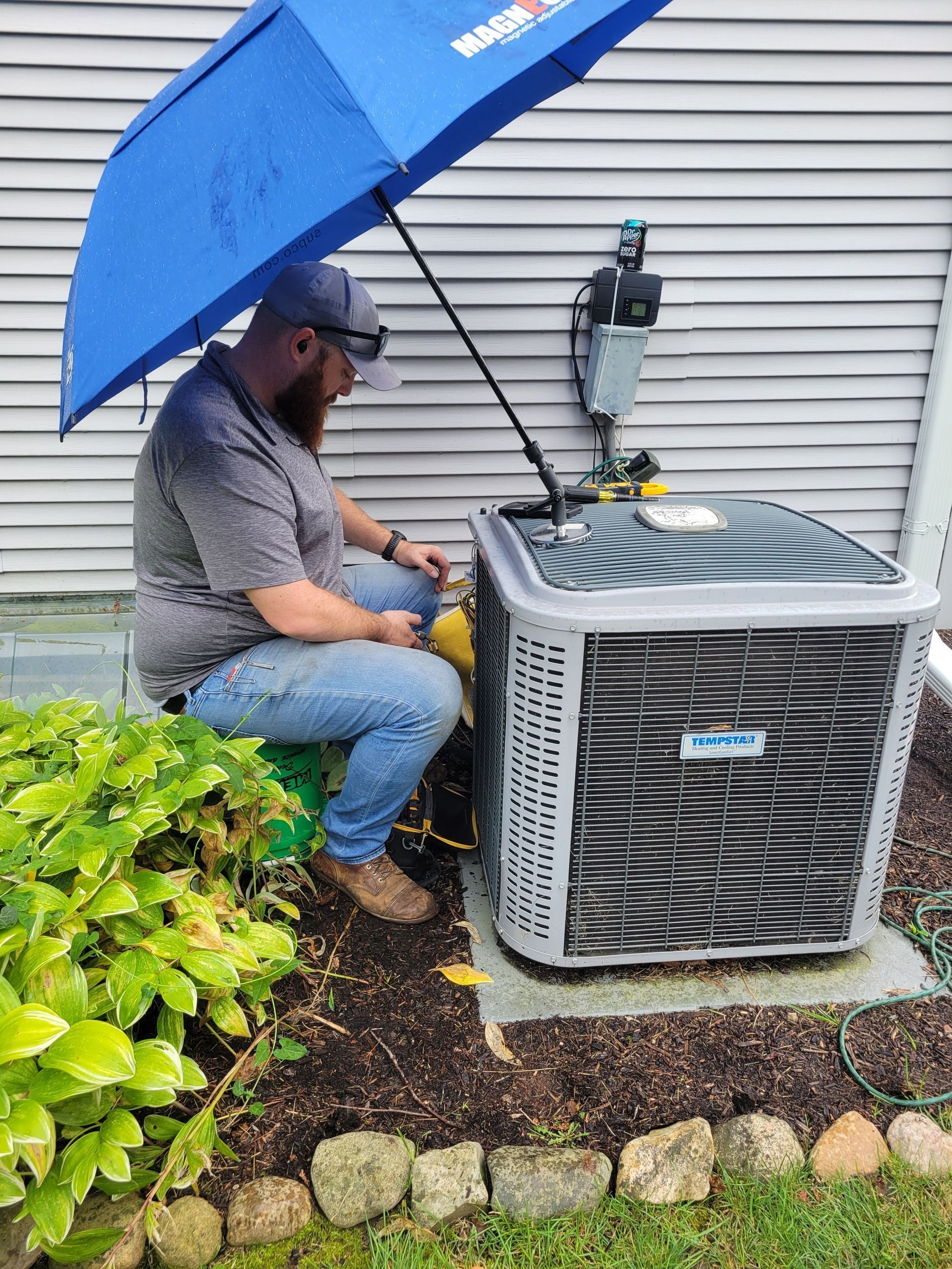 Technician under blue umbrella servicing outdoor air conditioner beside house siding