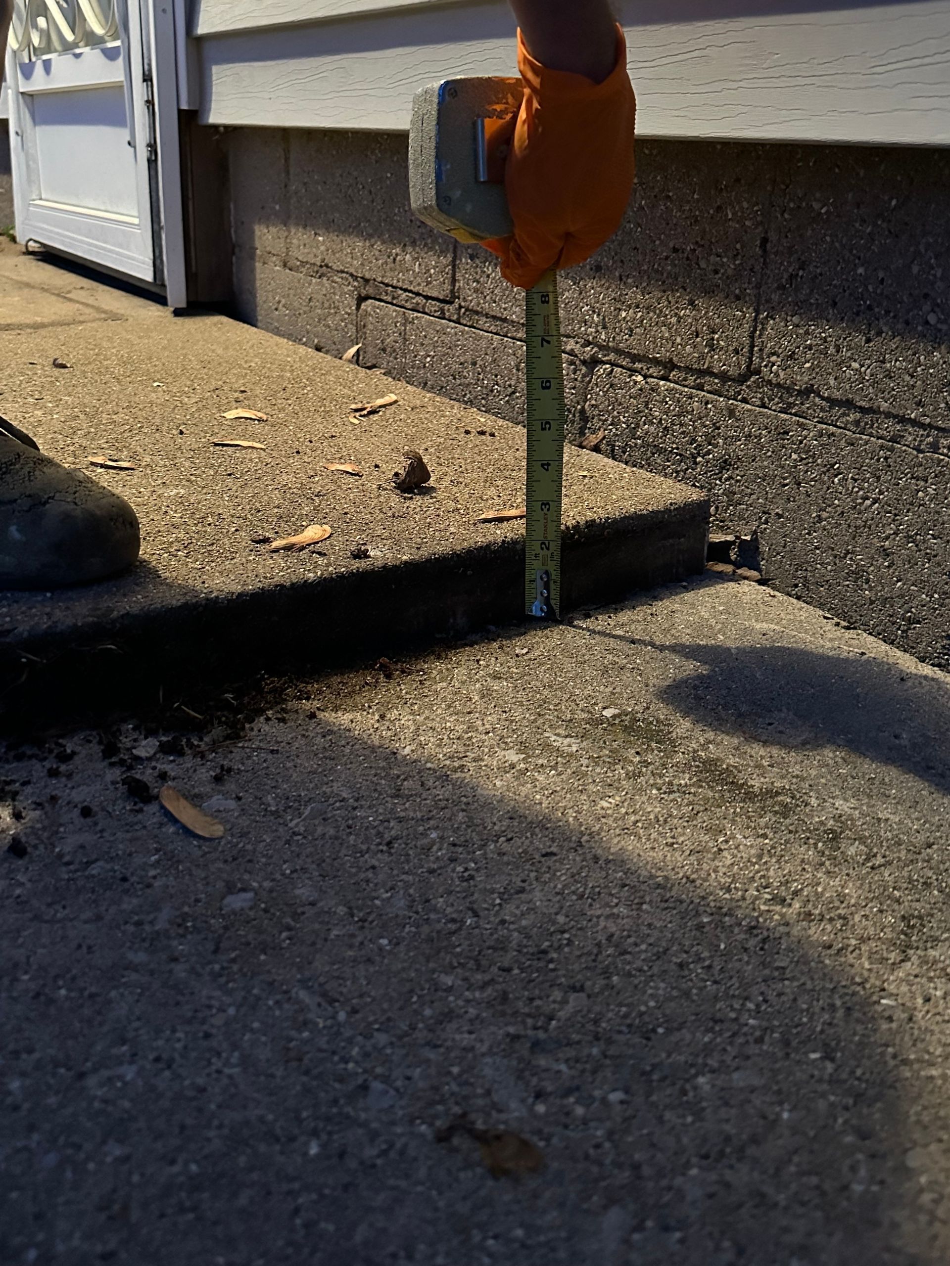 A gloved hand uses a tape measure to show a three-inch gap between a raised concrete step and the ground.