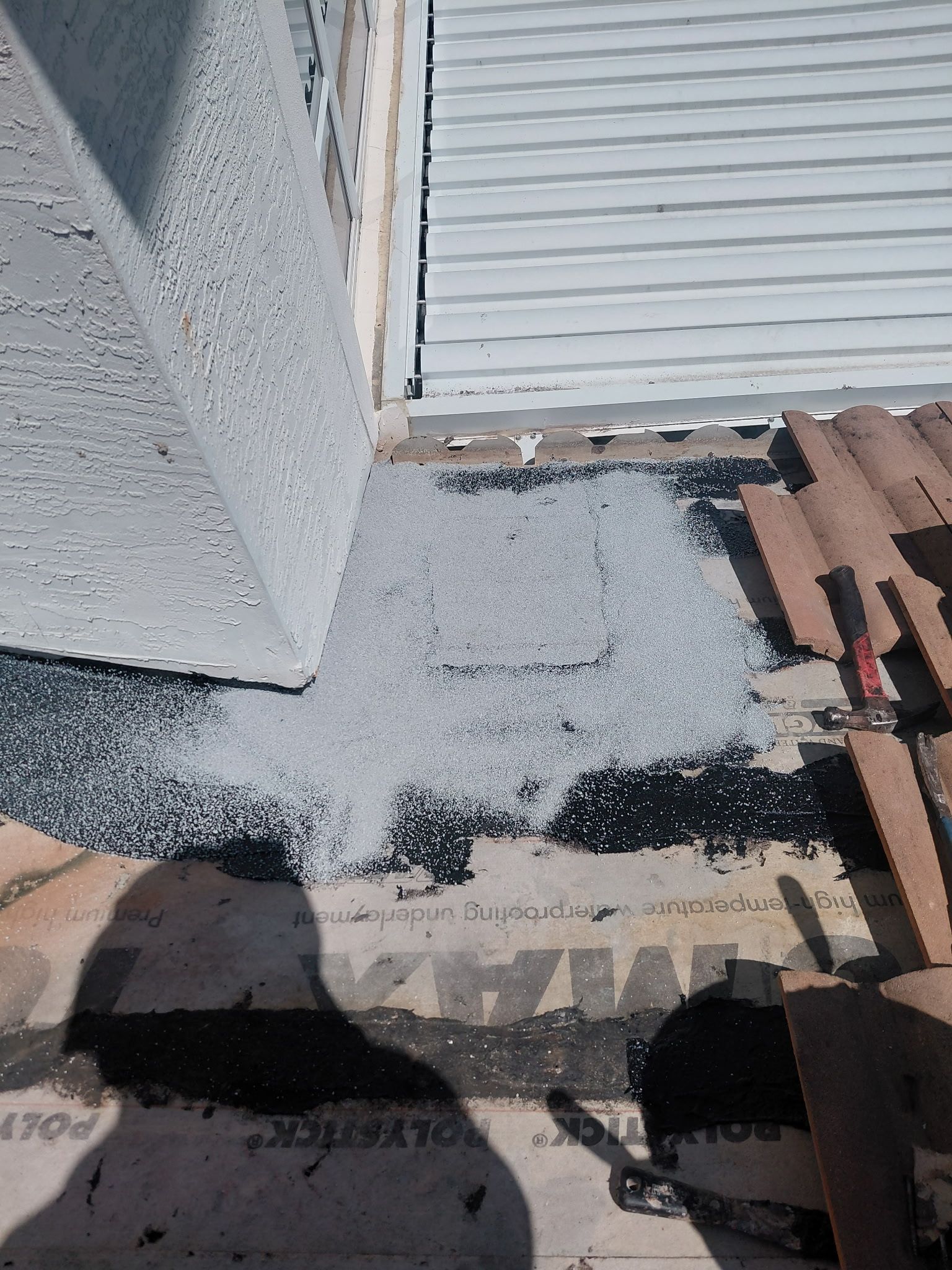 Roof with a damaged patch of white granules and exposed black material near a wall and gutter.