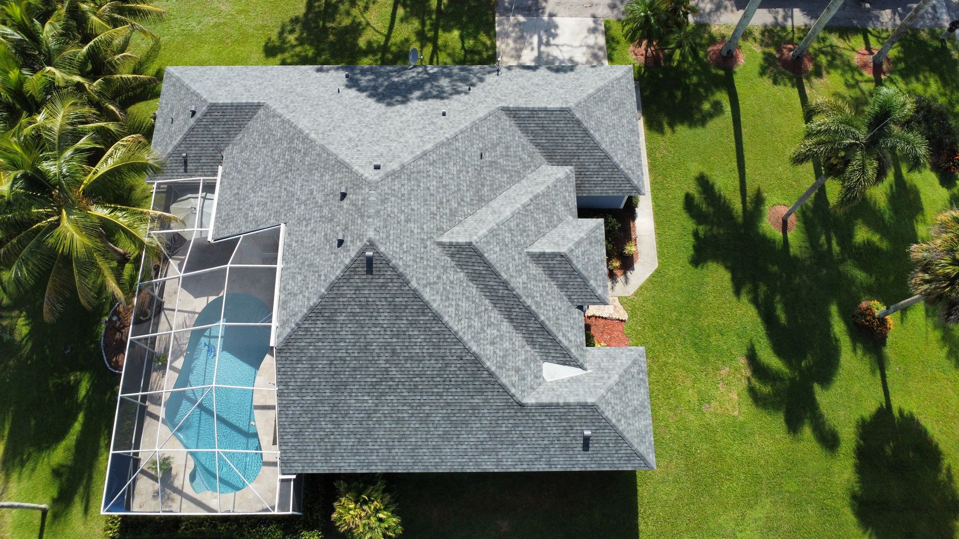 Aerial view of a house with a gray roof, screened pool enclosure, and green yard with palm trees.
