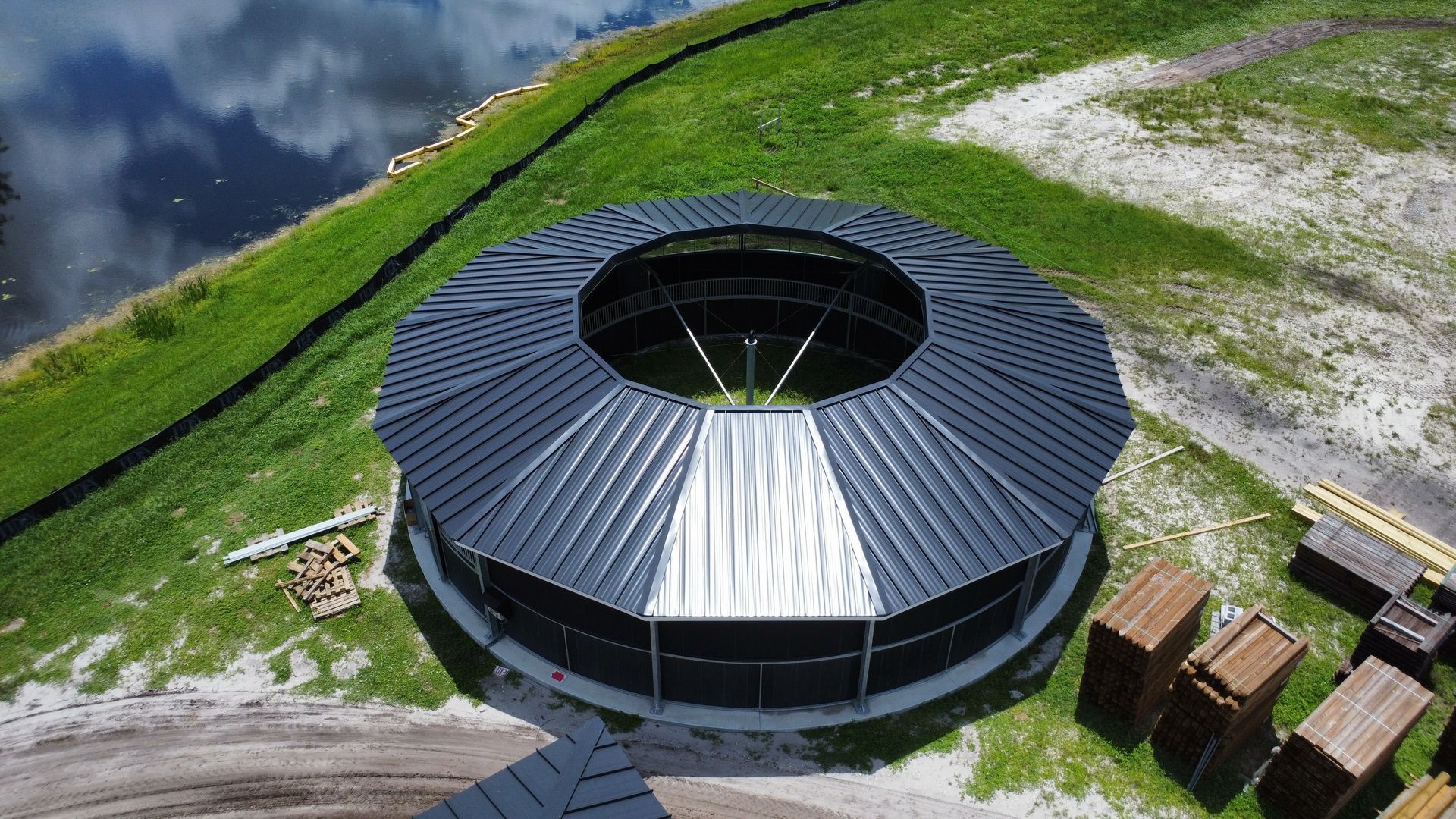 Aerial view of a round black-roofed building on a grassy mountainside near water and rocks