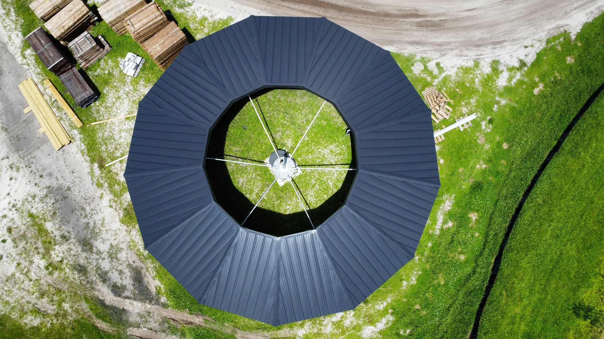 Aerial view of a circular dark-roofed building beside grass and stacked wooden pallets.