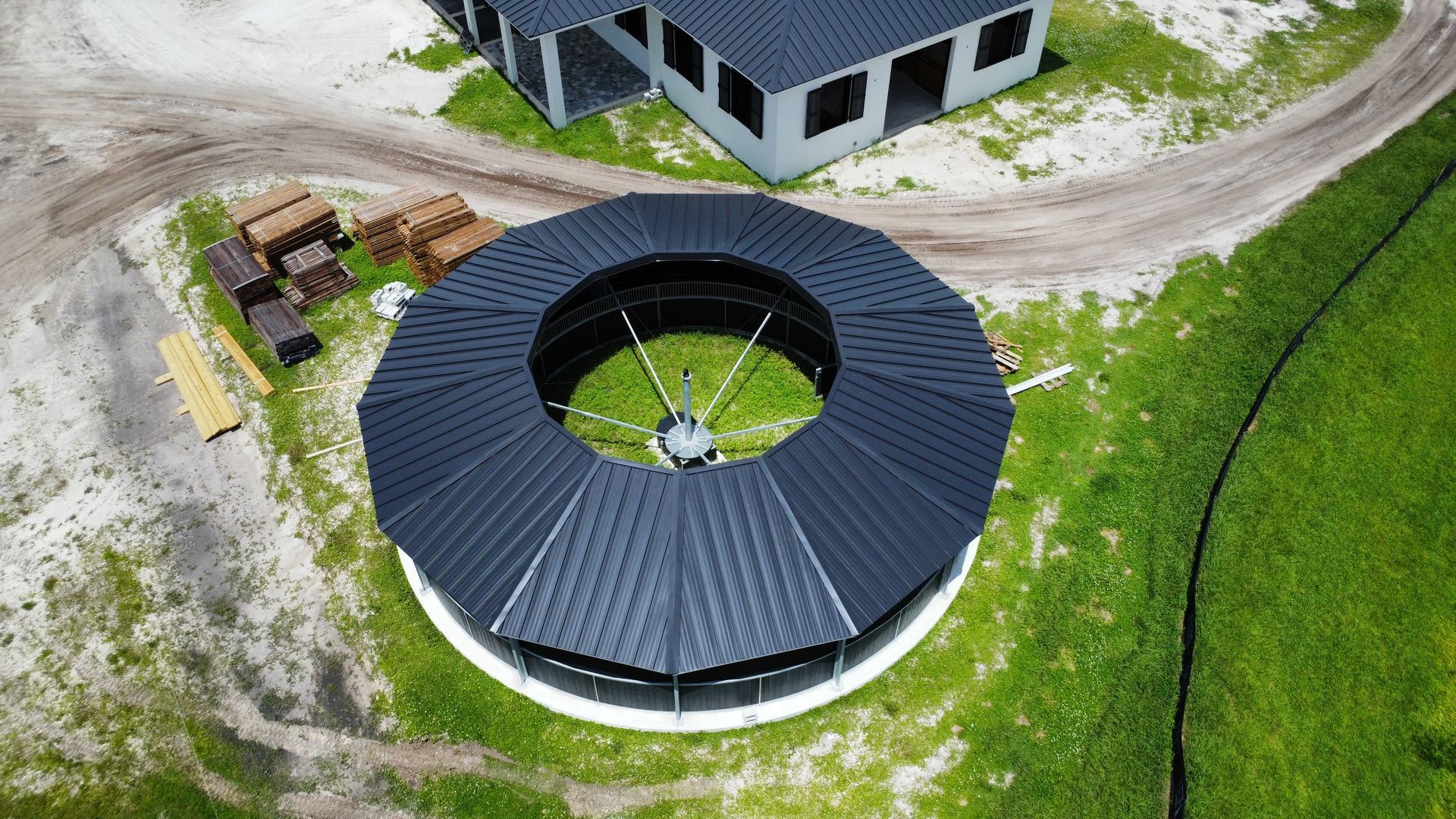 Aerial view of a circular building with a dark roof and central courtyard beside a dirt road and grassy field