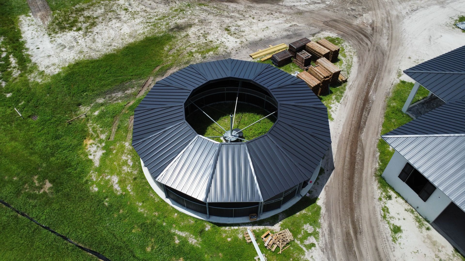 Aerial view of a circular black-roofed building beside dirt roads and green grass.