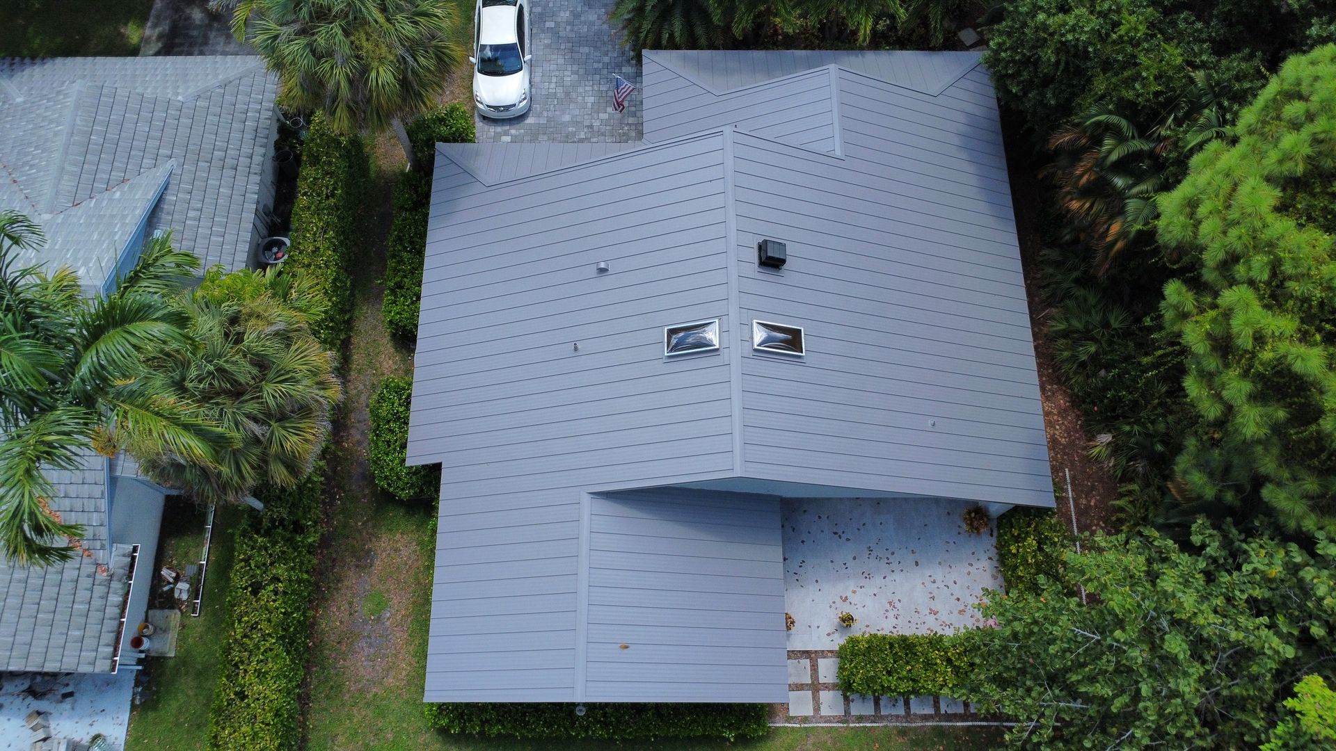 Aerial view of a gray-roofed house surrounded by dense green trees and a driveway with a parked car.