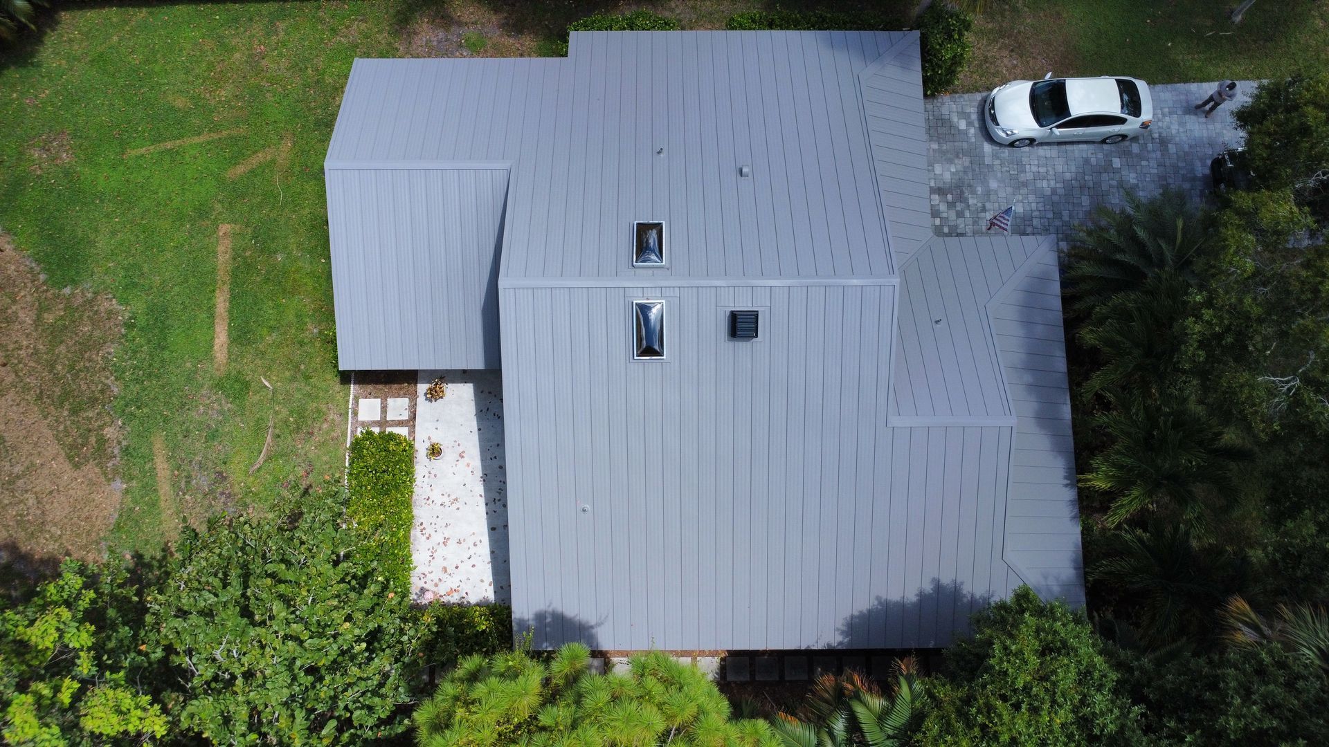 Aerial view of a gray-roofed house with a driveway and parked car, surrounded by green trees.