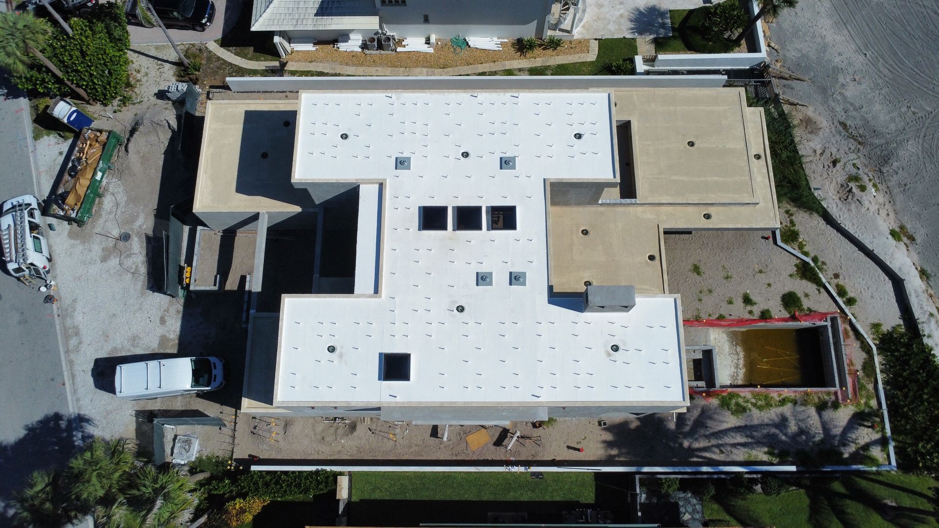 Aerial view of a large white-roofed building with a beige wing, parked vehicles, and surrounding roads.