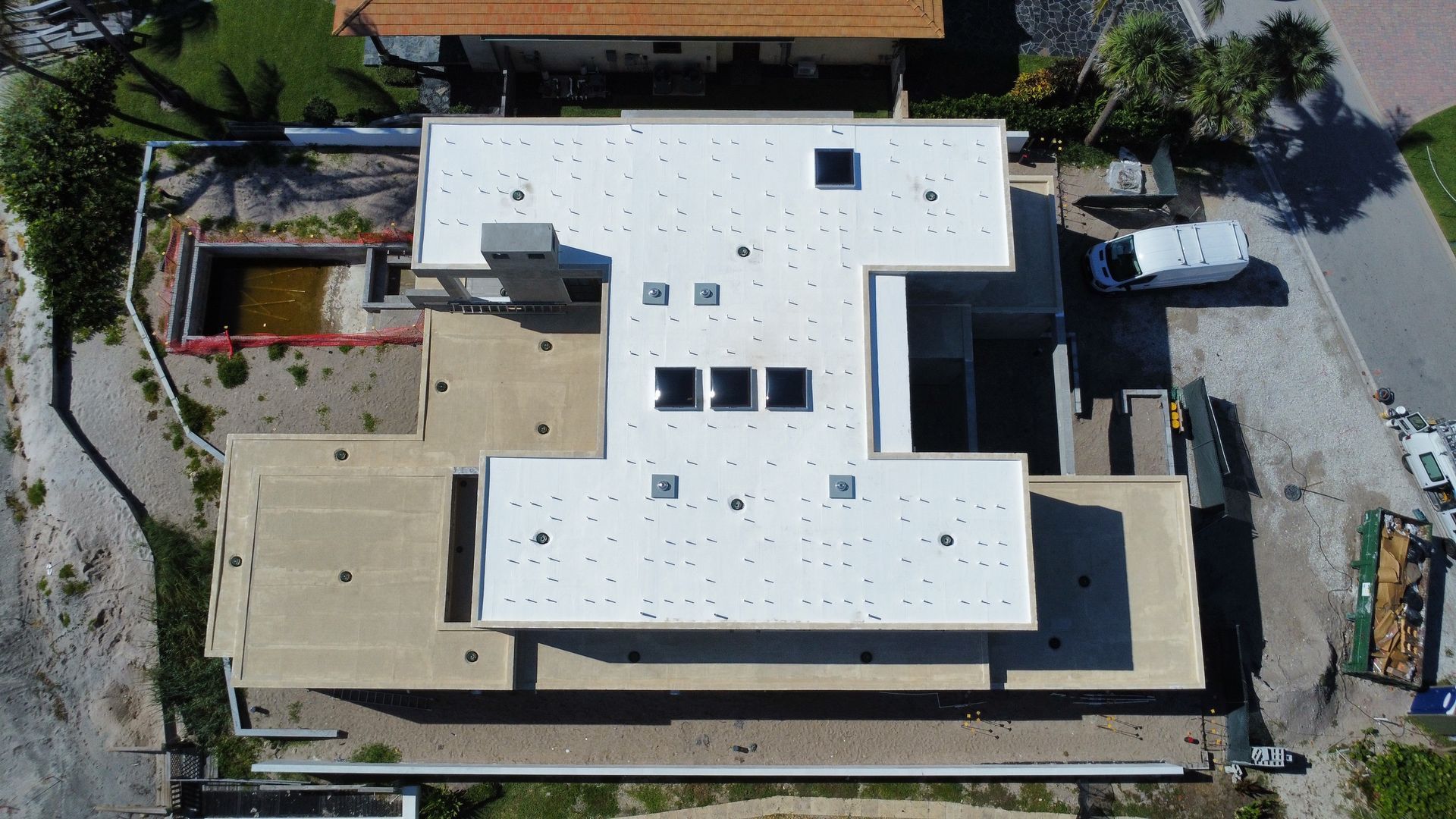 Aerial view of a white-roofed L-shaped building surrounded by trees and paved areas.