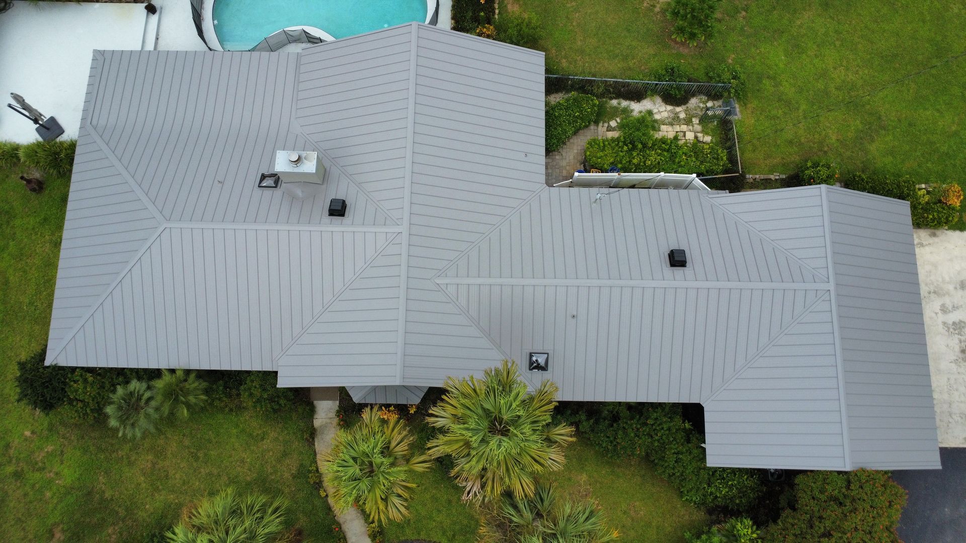 Aerial view of a gray shingled house roof with vents, surrounded by green lawn and trees.