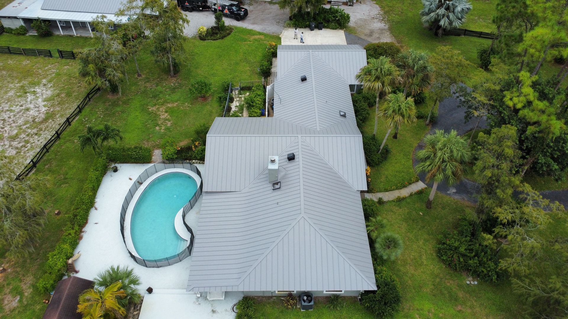 Aerial view of a house with a light gray roof and a curved backyard swimming pool surrounded by greenery