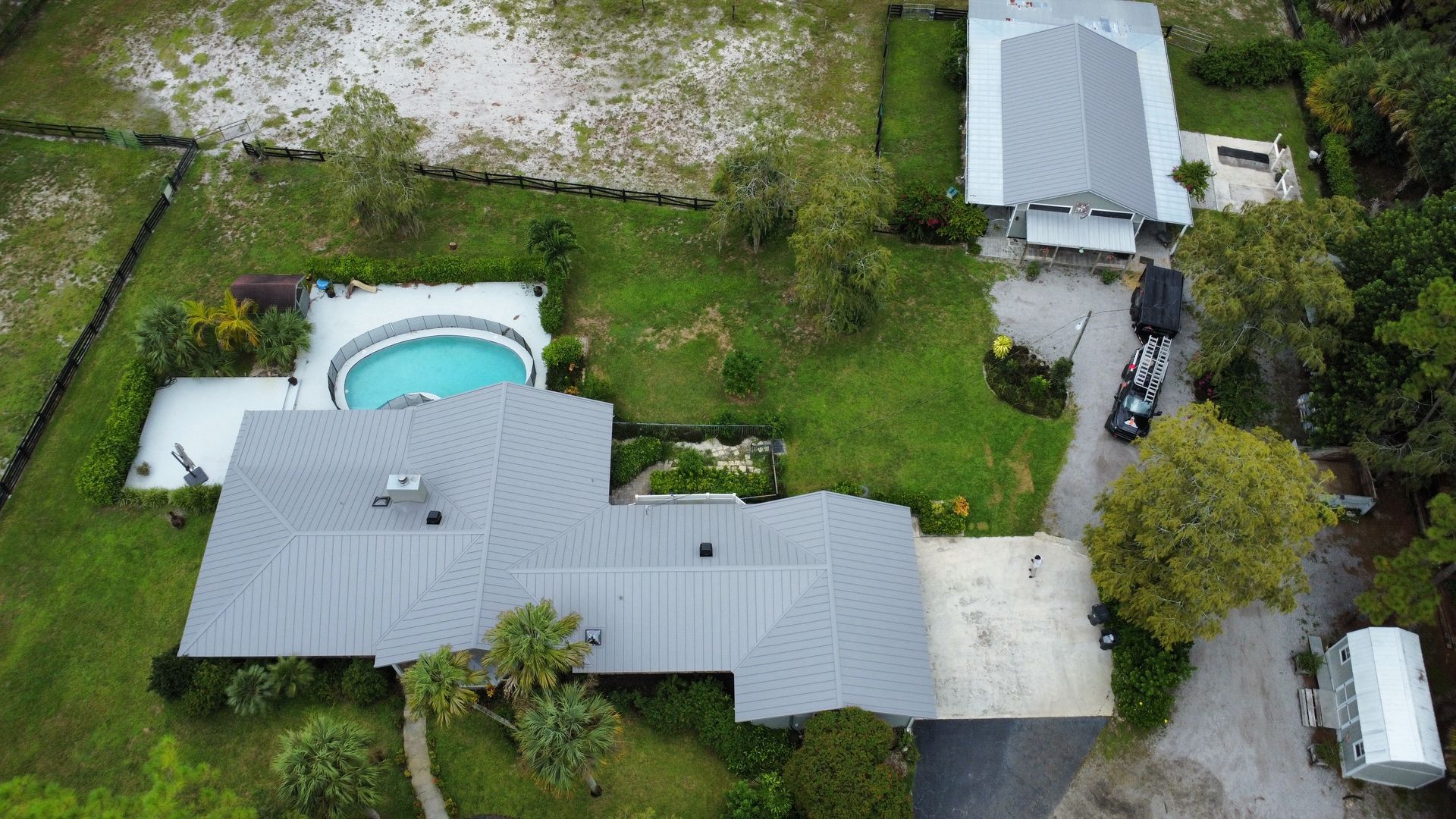 Aerial view of a house with a backyard pool, driveway, and surrounding trees and lawn.