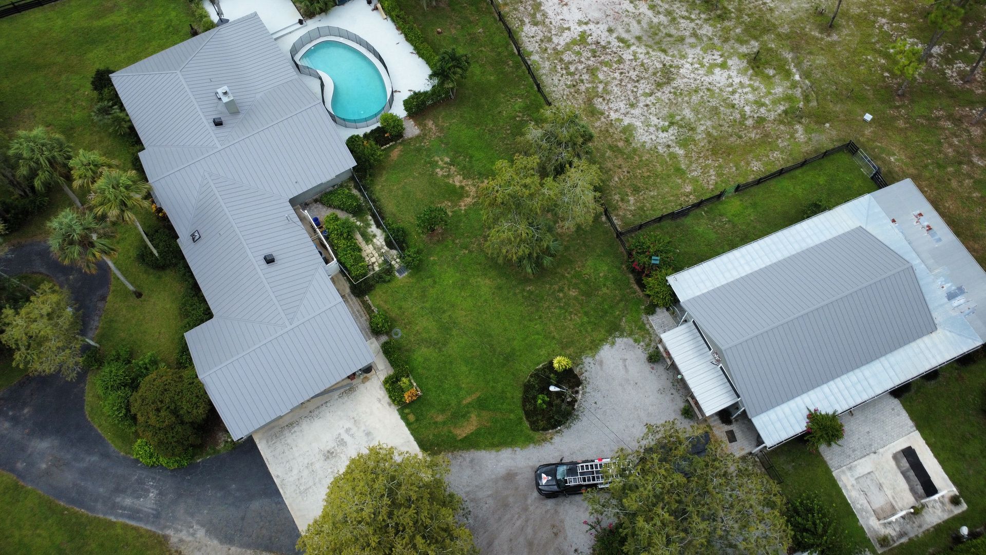Aerial view of two houses with gray roofs, a backyard pool, driveway, and surrounding green yard.