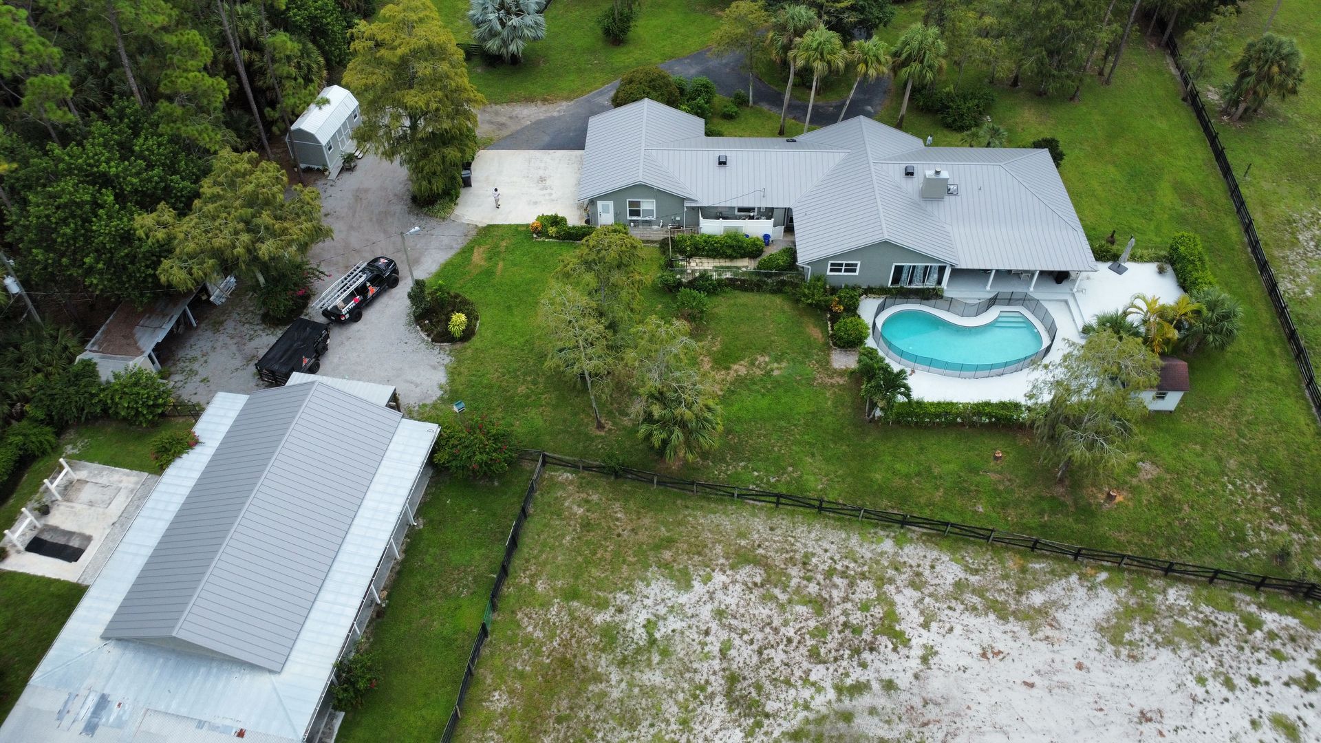 Aerial view of a house with a backyard pool, driveway, and detached garage surrounded by trees and grass.