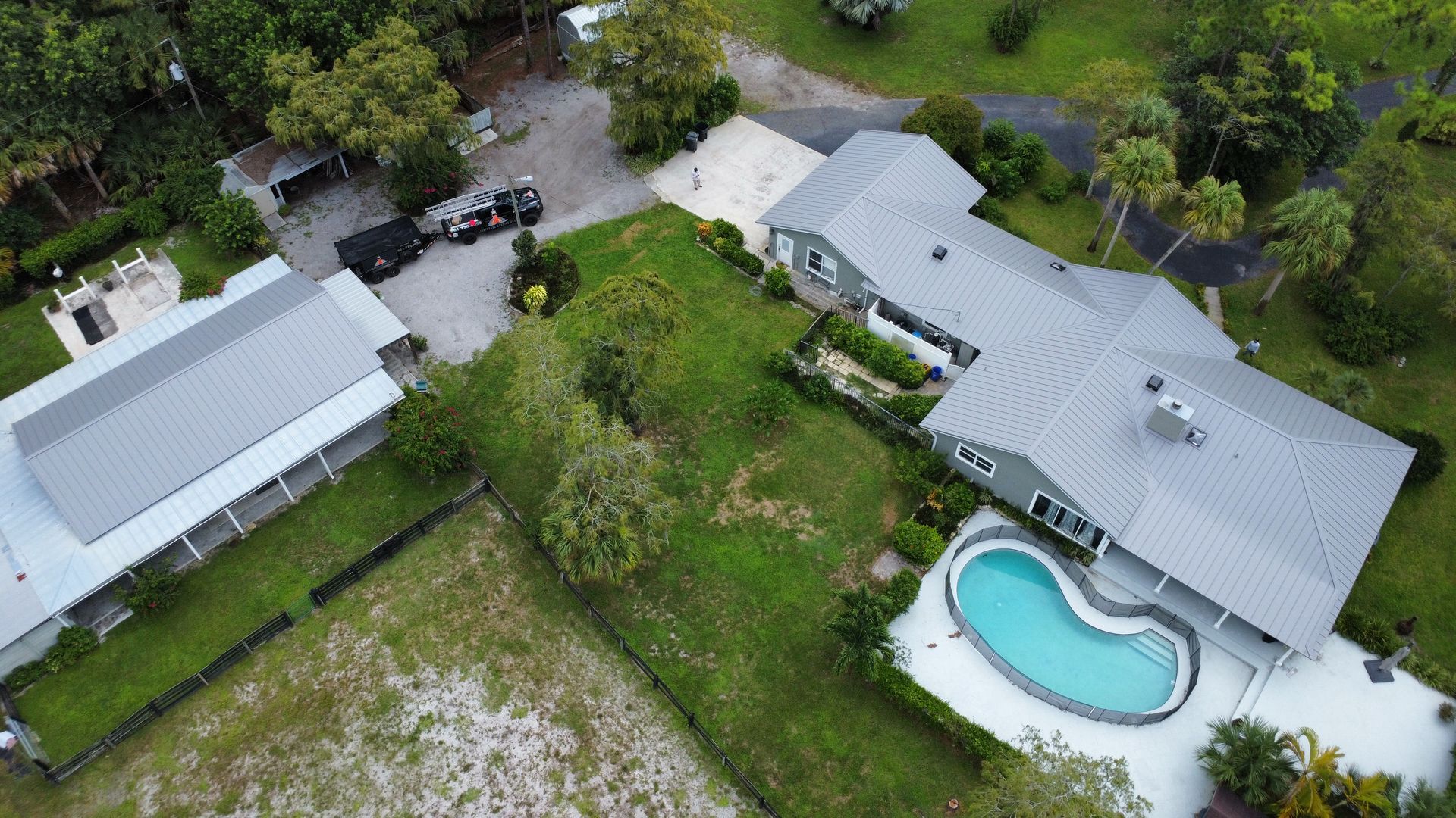 Aerial view of a house with a turquoise pool, driveway, and green yard surrounded by trees.