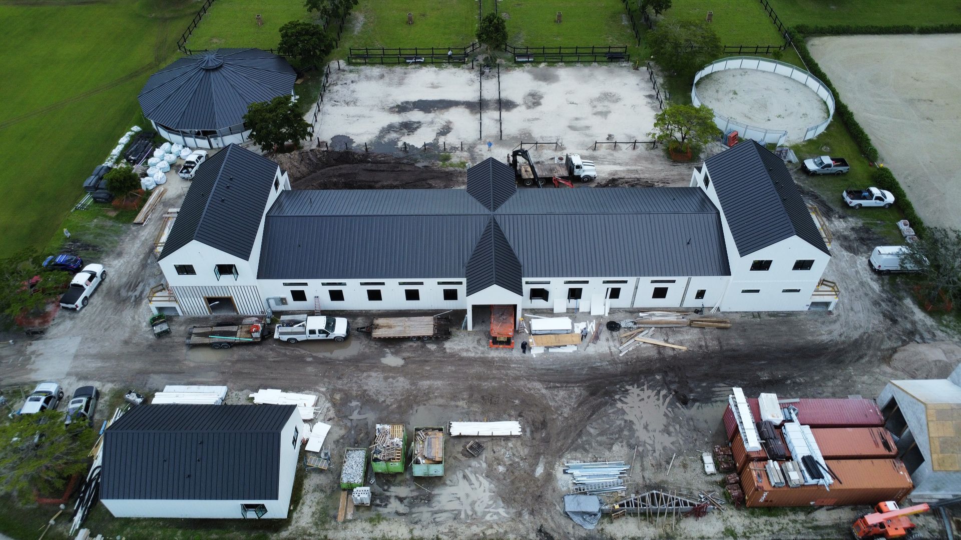 Aerial view of a long white building with a dark roof and two round tanks, surrounded by pavement and grass.