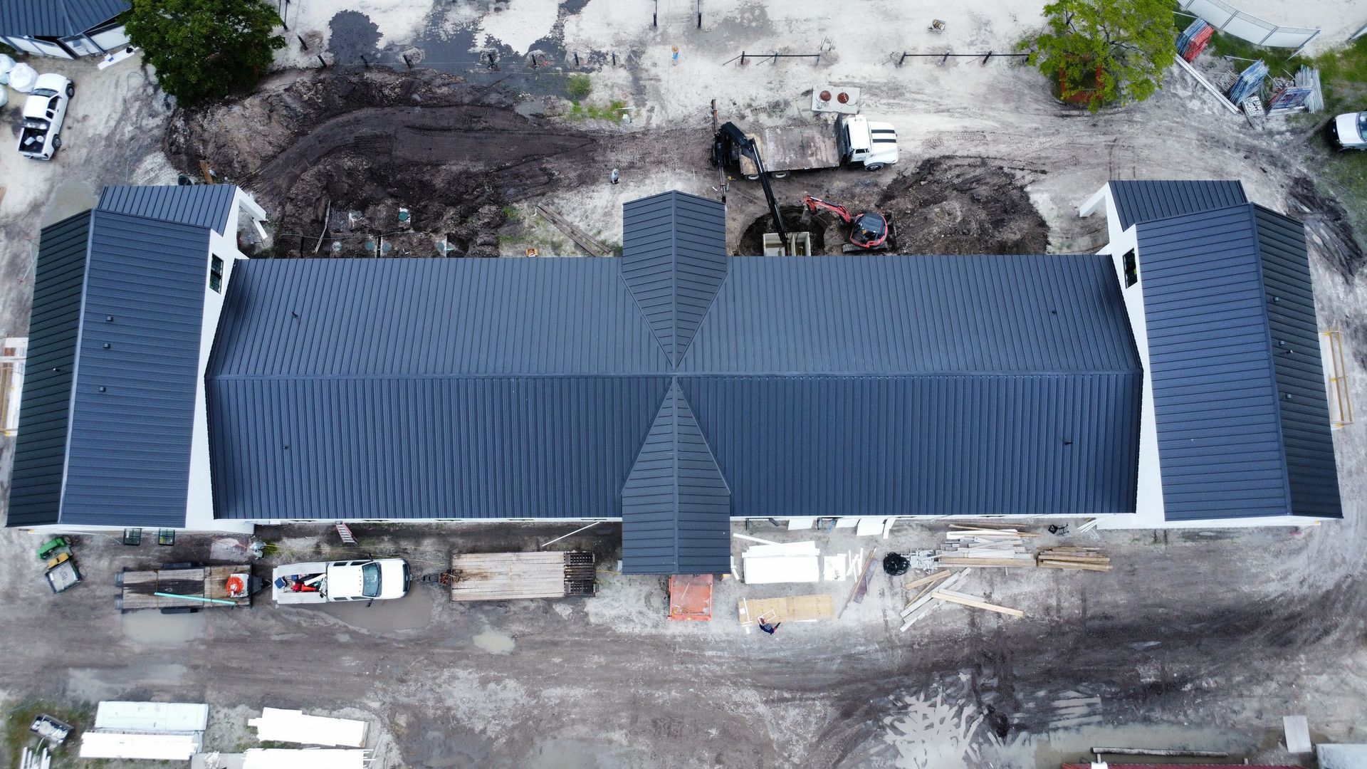 Aerial view of a long blue-roofed building with side wings, surrounded by dirt, equipment, and trees.