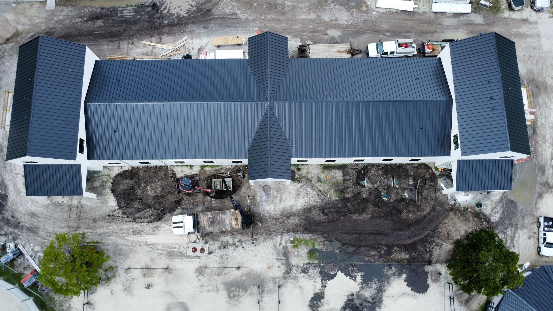 Aerial view of a long building with a dark blue roof and surrounding dirt lot and parked vehicles