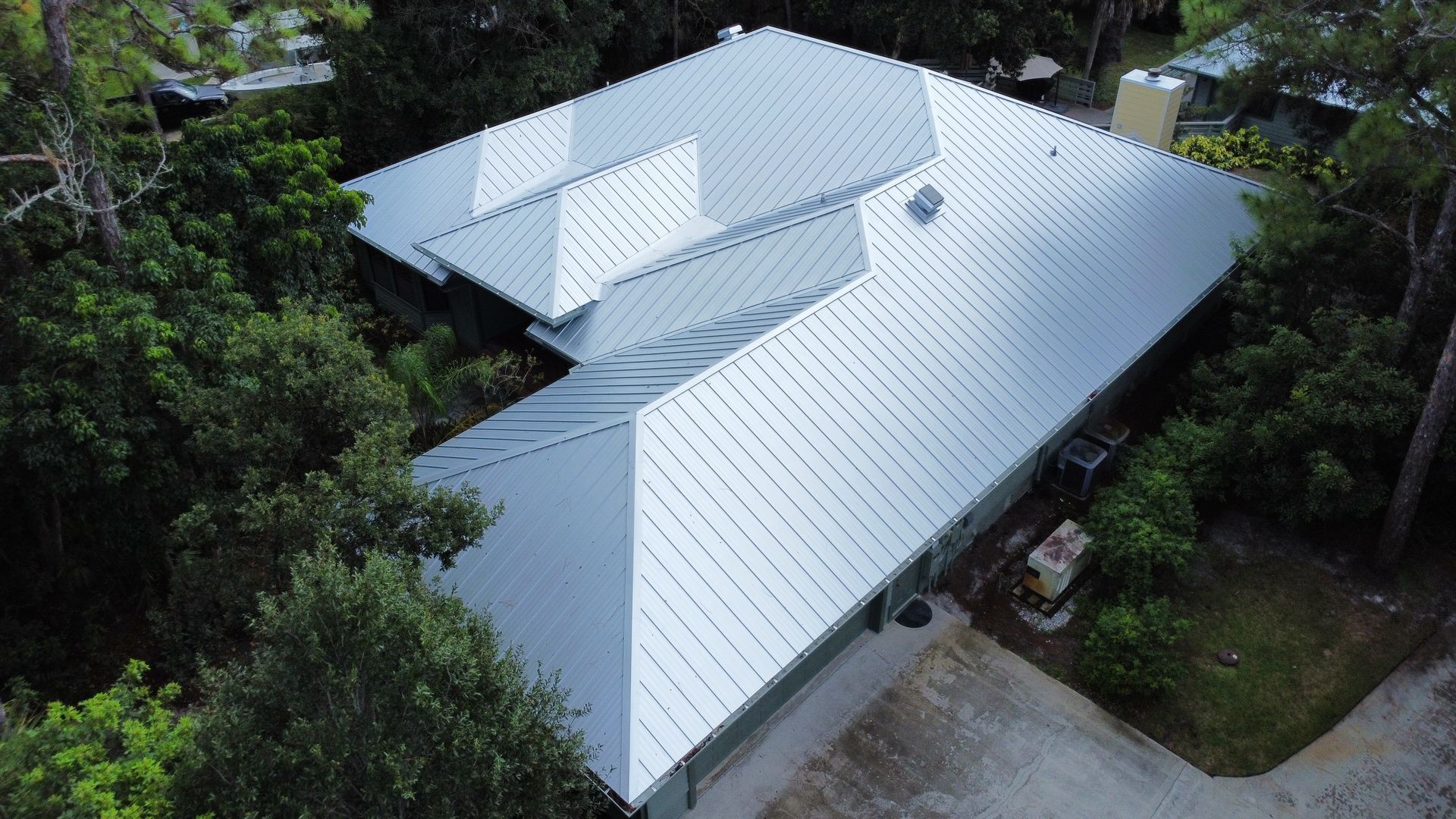 Aerial view of a house with a white metal roof surrounded by trees and a driveway.