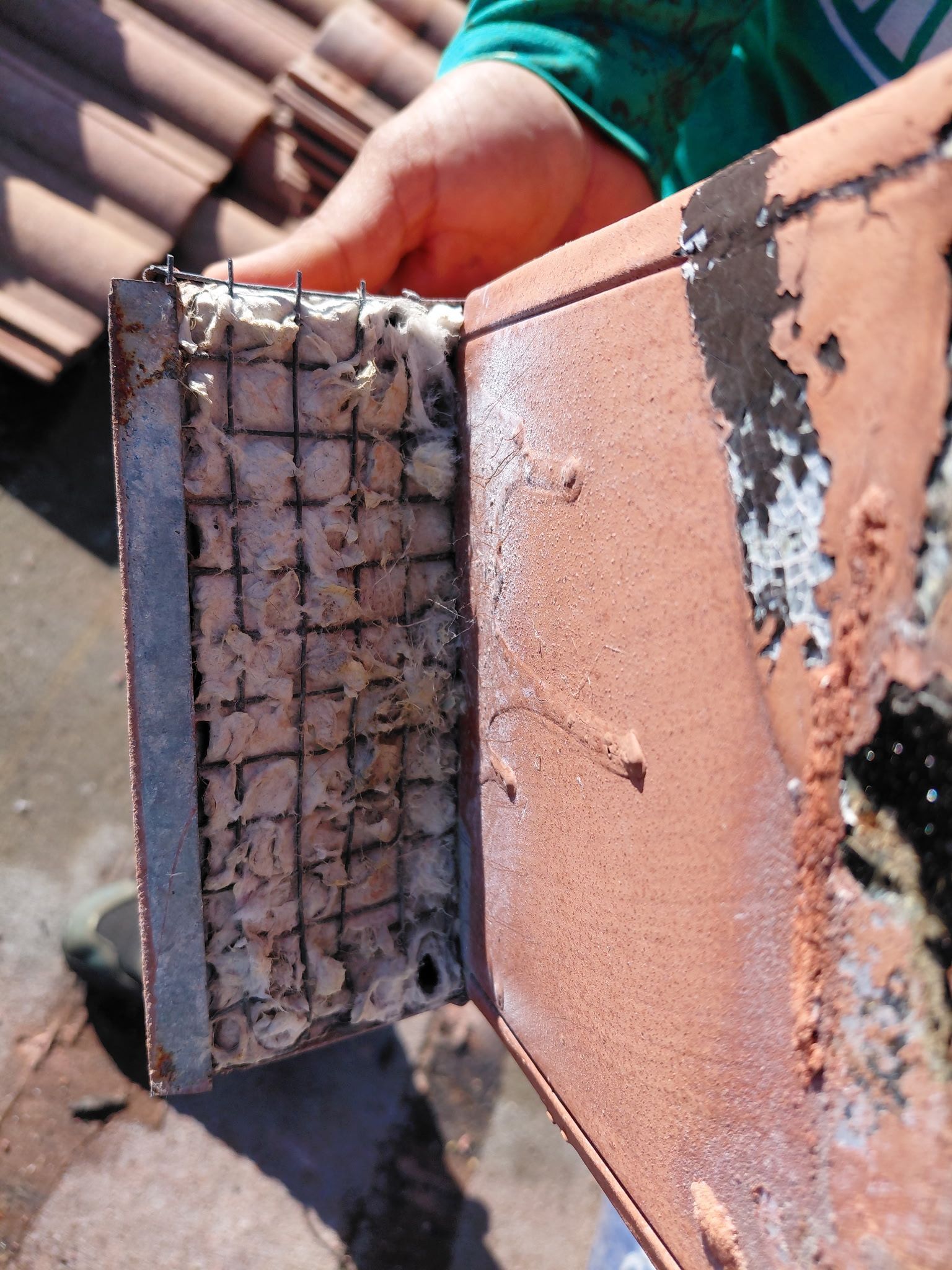 Close-up of a damaged roof tile with exposed mesh and cracked stucco, held by a hand outdoors.