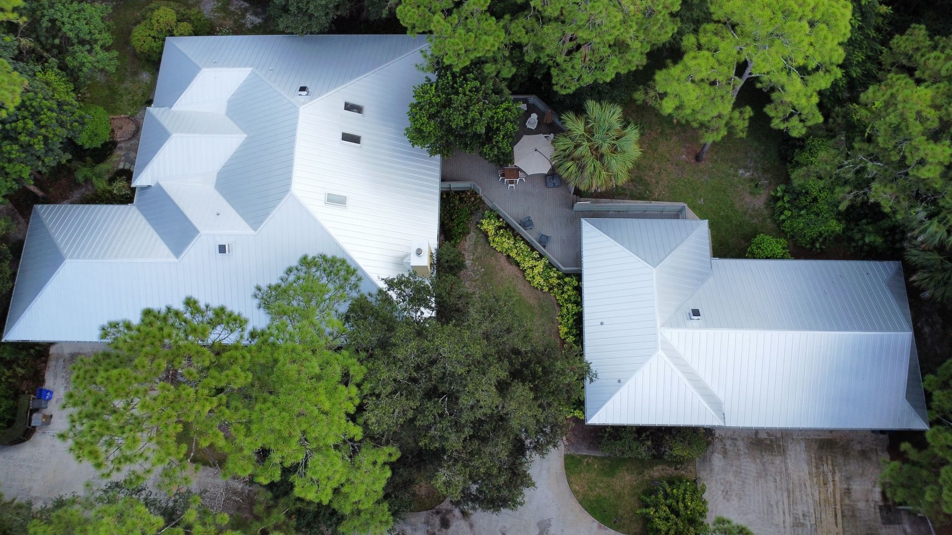 Aerial view of two light-roofed houses surrounded by dense green trees and a driveway.