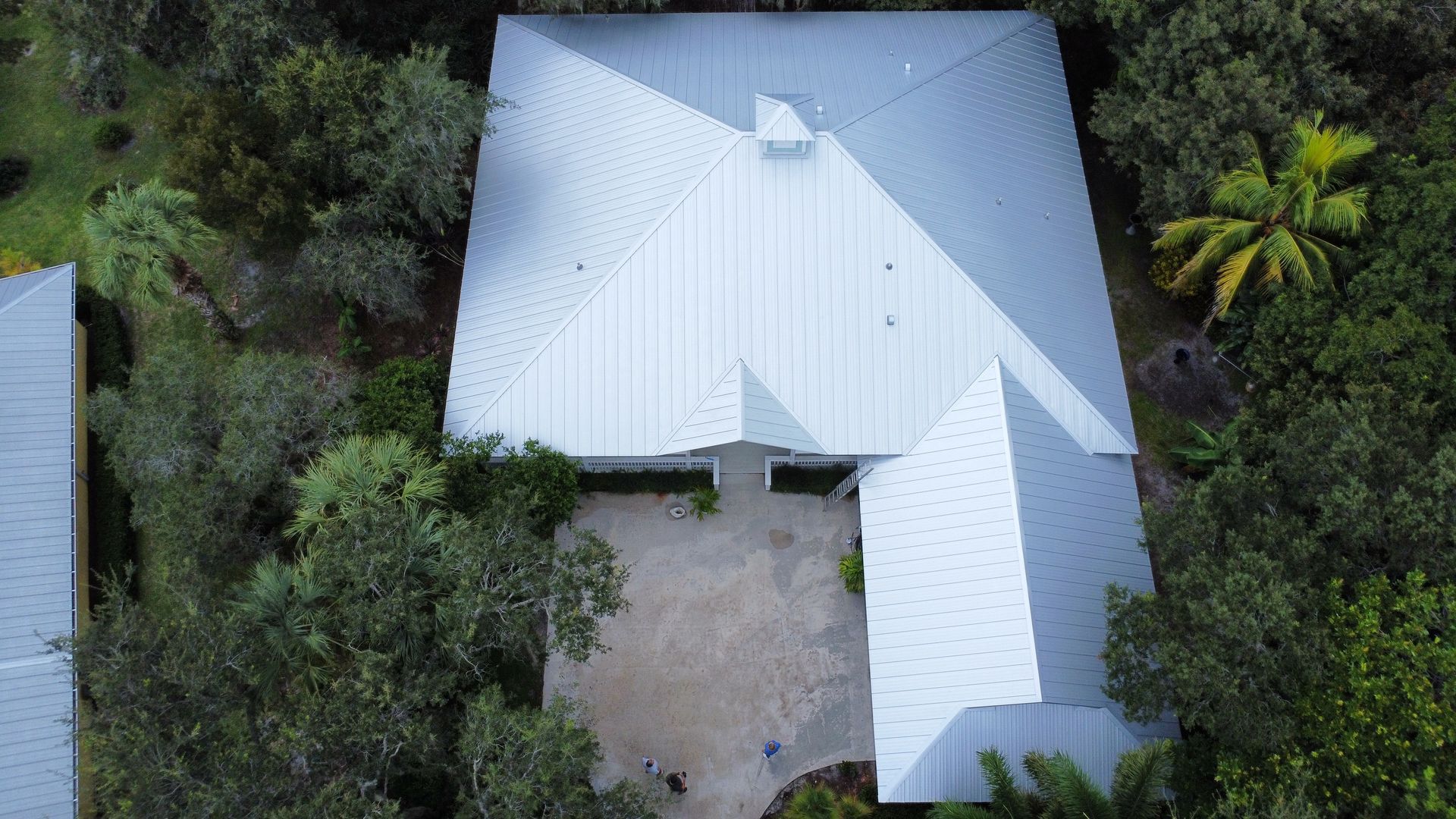 Aerial view of a light gray metal-roofed house surrounded by trees and a small courtyard.