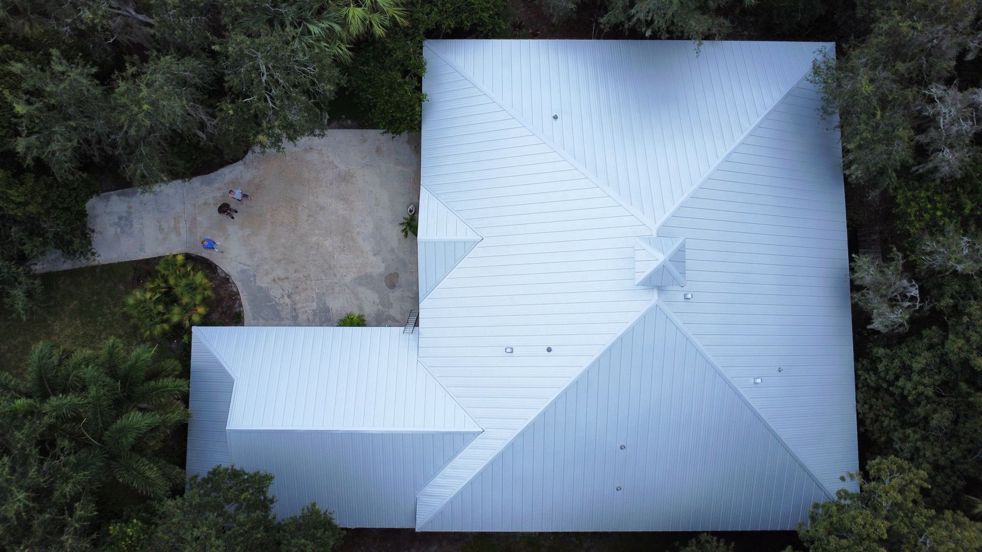 Aerial view of a white-roofed house surrounded by dense green trees and a driveway on the left.