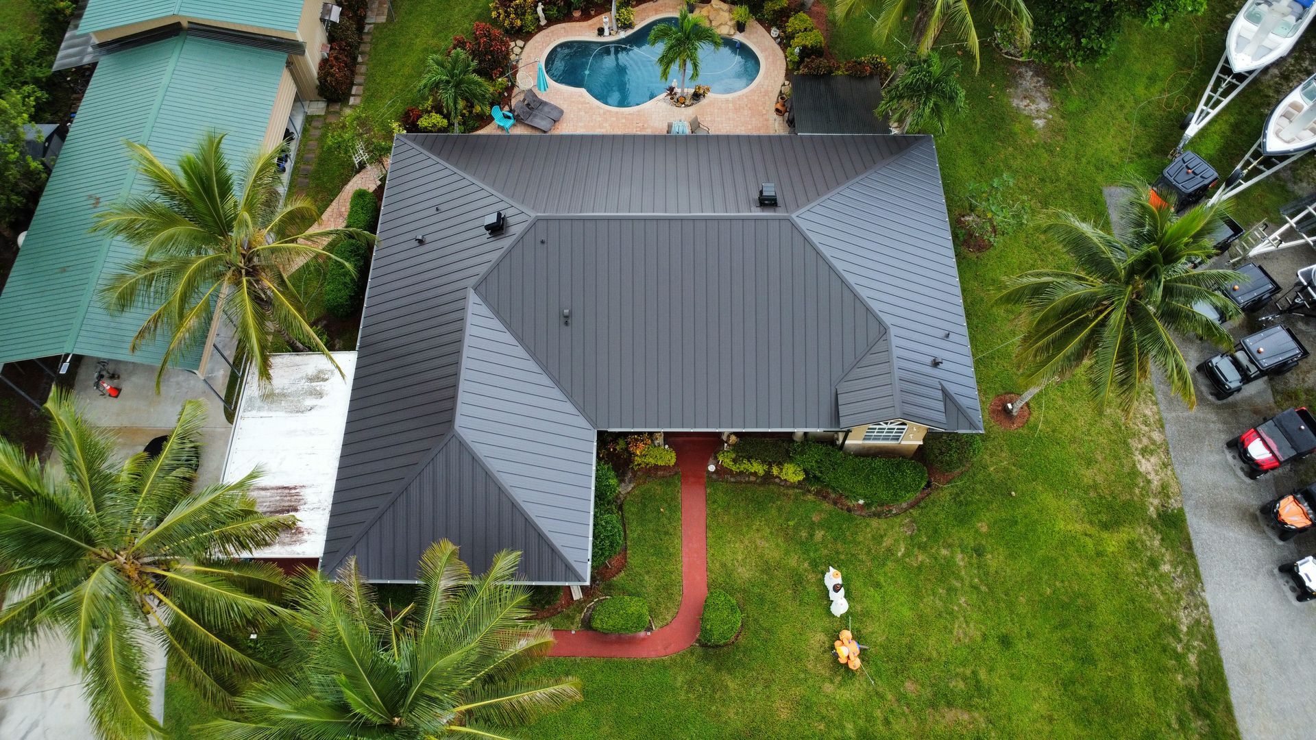 Aerial view of a gray-roofed house with a backyard pool, lawn, palm trees, and parked cars nearby