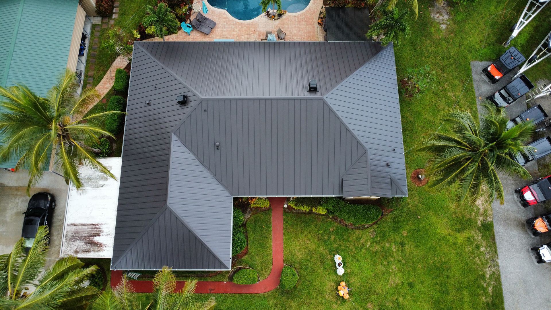 Aerial view of a gray-roofed house with a red walkway, surrounded by green lawn and palm trees