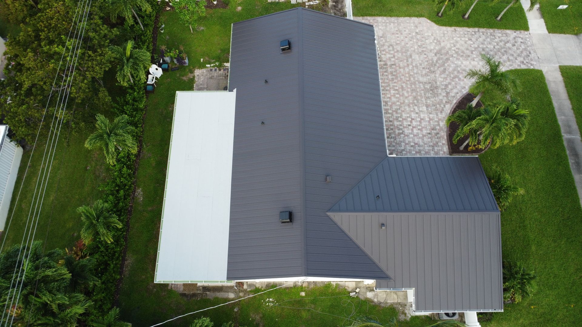 Aerial view of a house with dark gray roof, white extension, and surrounding green yard and driveway