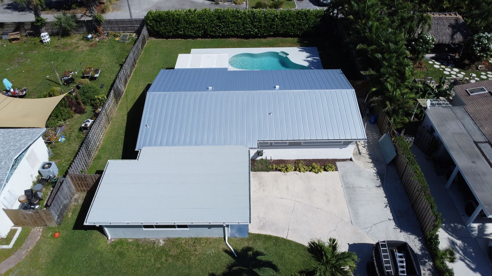 Aerial view of a white-roofed house with a swimming pool, lawn, and surrounding trees.