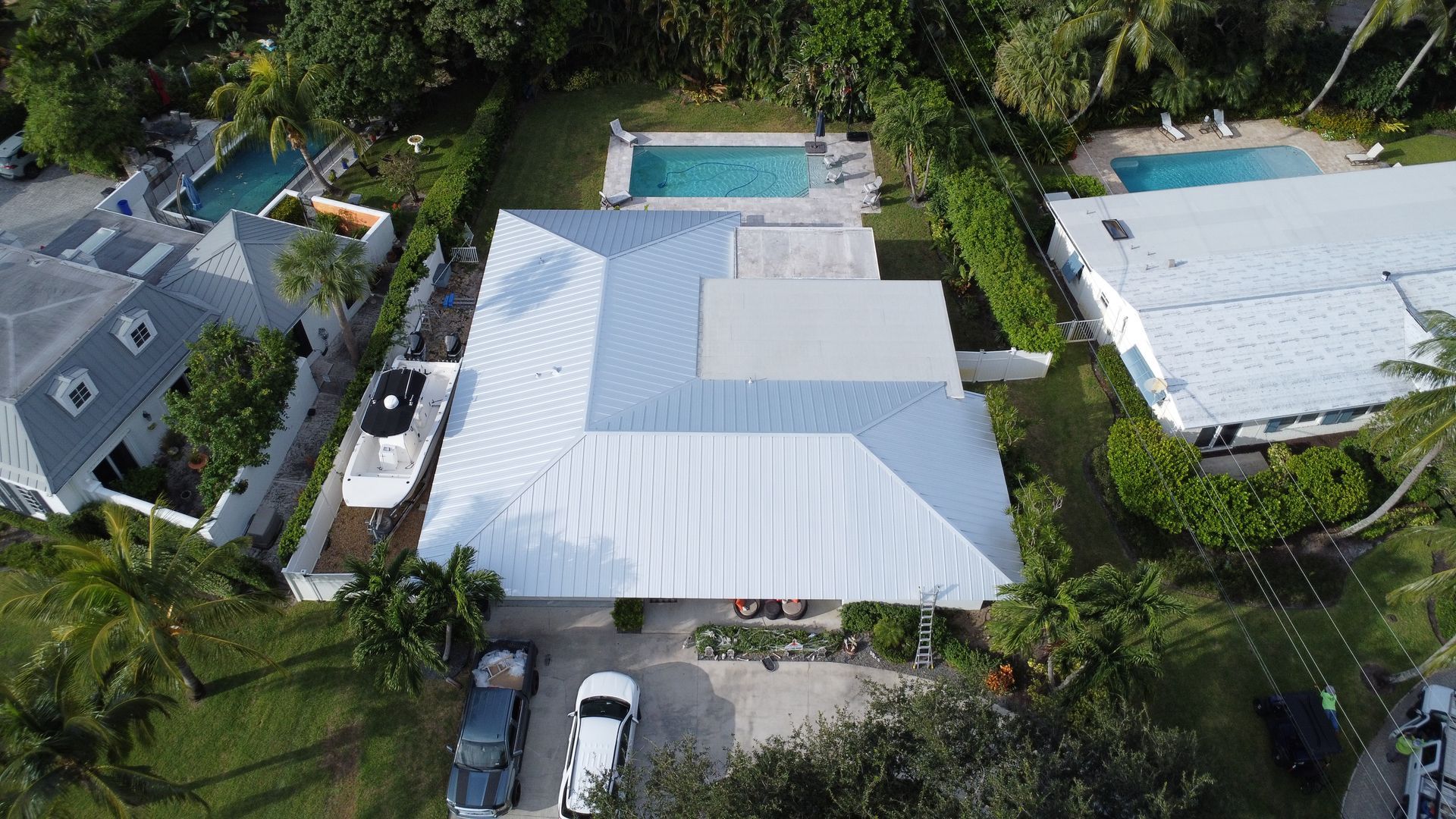 Aerial view of a white-roofed house with a backyard pool, surrounded by trees and neighboring homes.