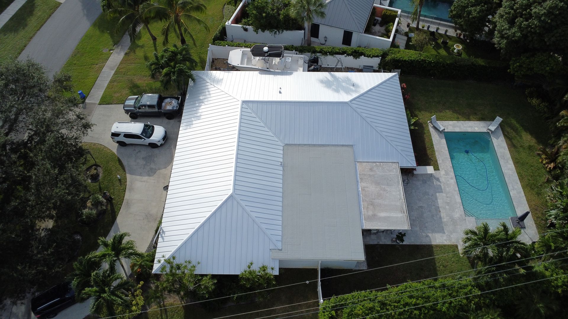 Aerial view of a white-roofed house with a driveway, parked cars, and a rectangular swimming pool.