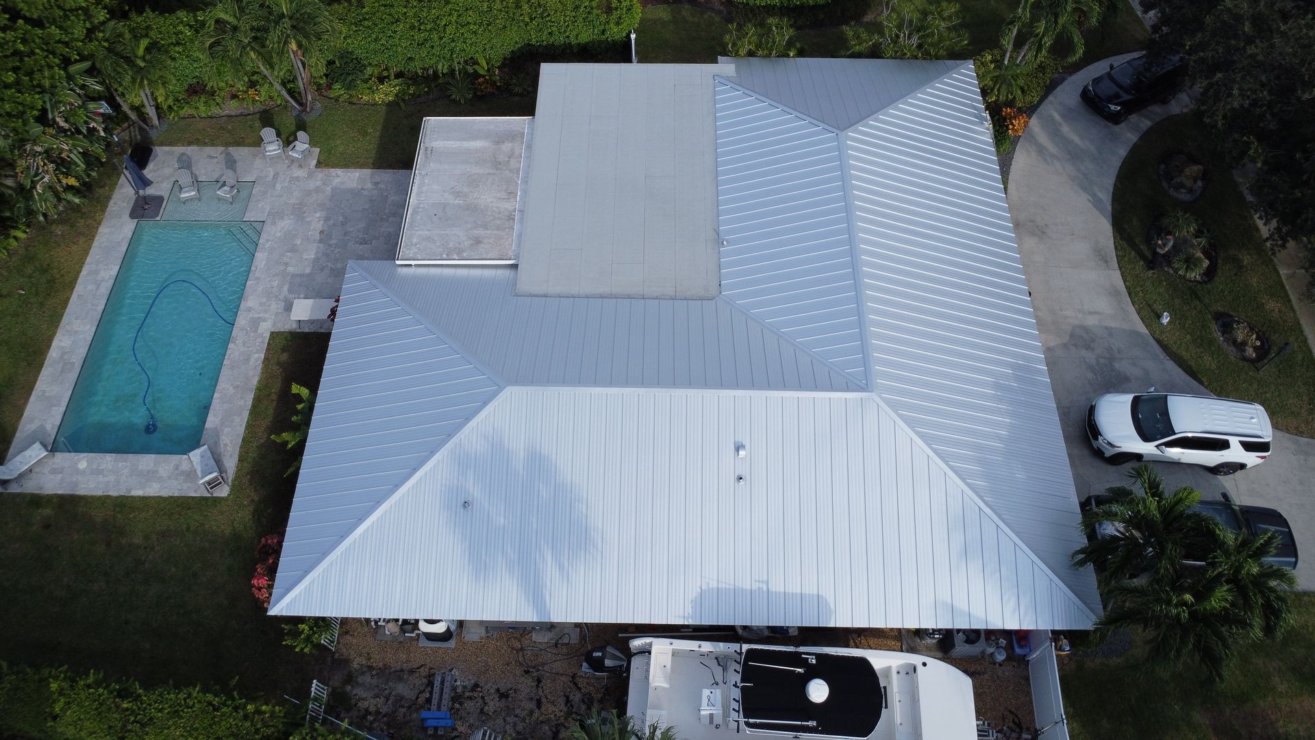 Aerial view of a house with a white metal roof, driveway, parked cars, and a swimming pool.