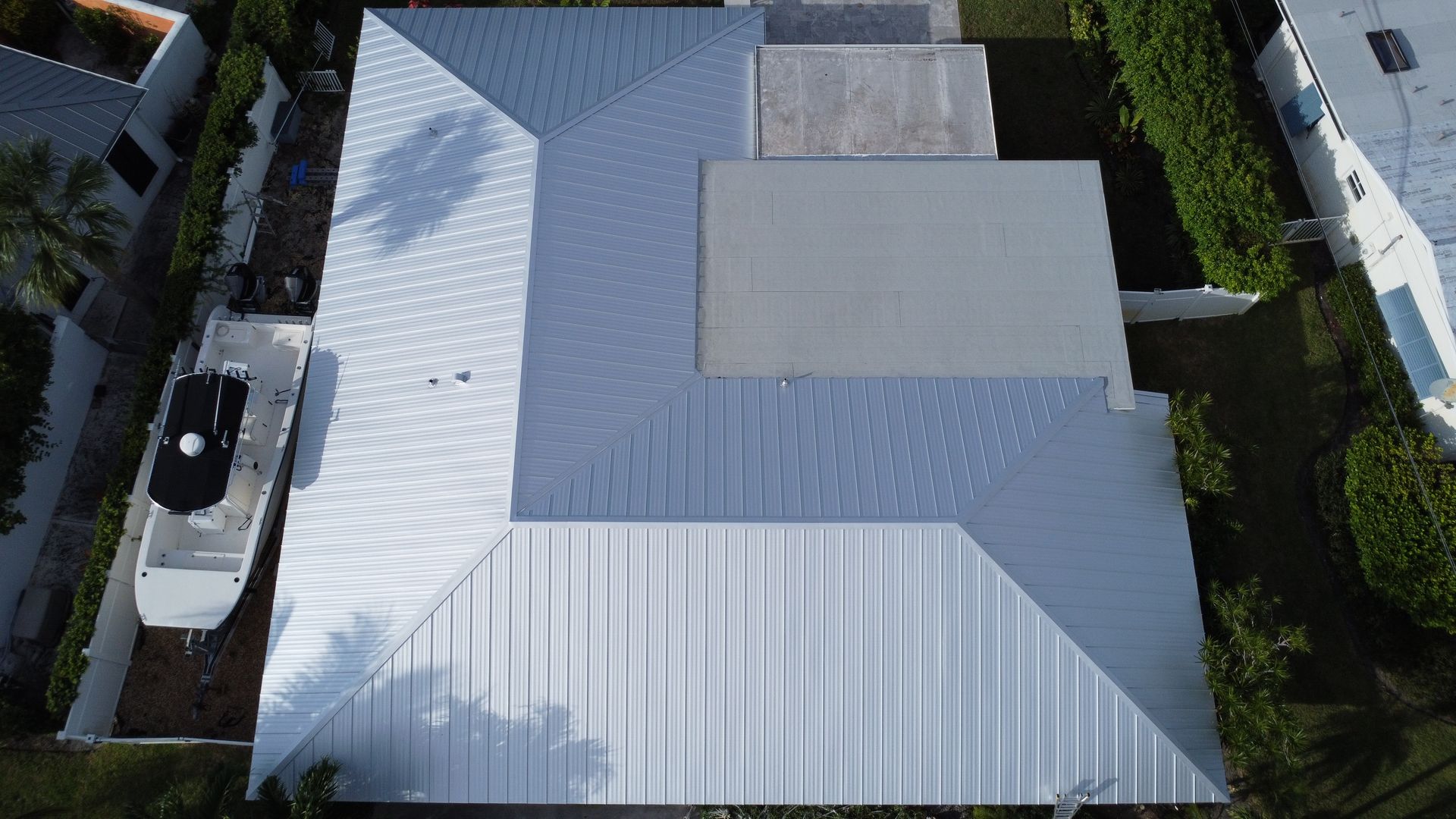 Aerial view of a white-roofed house with a central flat section, surrounded by trees and neighboring homes.